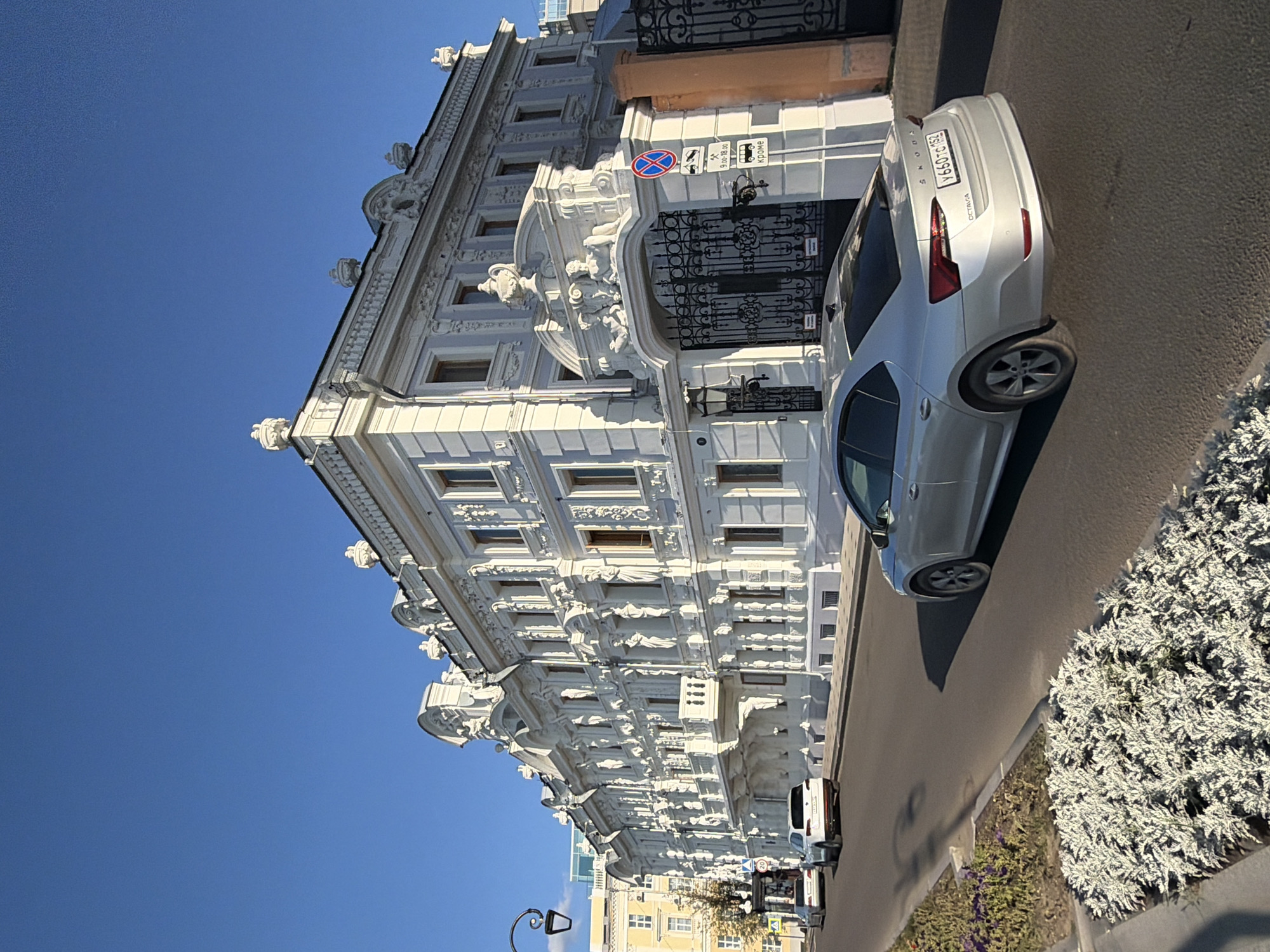 An outdoor, eye-level, wide shot captures a bright, sunny day in Nizhny Novgorod, Russia. The focus is on a grand, white, ornate building with intricate architectural details, including statues, carvings, and a balcony. The building features multiple stories and numerous windows. To the right of the building is a large, black, wrought-iron gate, topped with decorative elements. A red and blue circular sign with a diagonal red bar and a blue prohibition symbol is posted on a pole next to the gate, along with other signage. A silver Skoda Octavia sedan is parked on the right side of the road, its rear facing the camera. The car has a license plate that reads "Y660TC 152". In the background, other cars are visible on the street, and a portion of a yellow building can be seen to the left. The street is lined with a sidewalk and a planter box filled with lush, silver-grey foliage, with some purple flowers interspersed. The sky is a clear, vibrant blue.