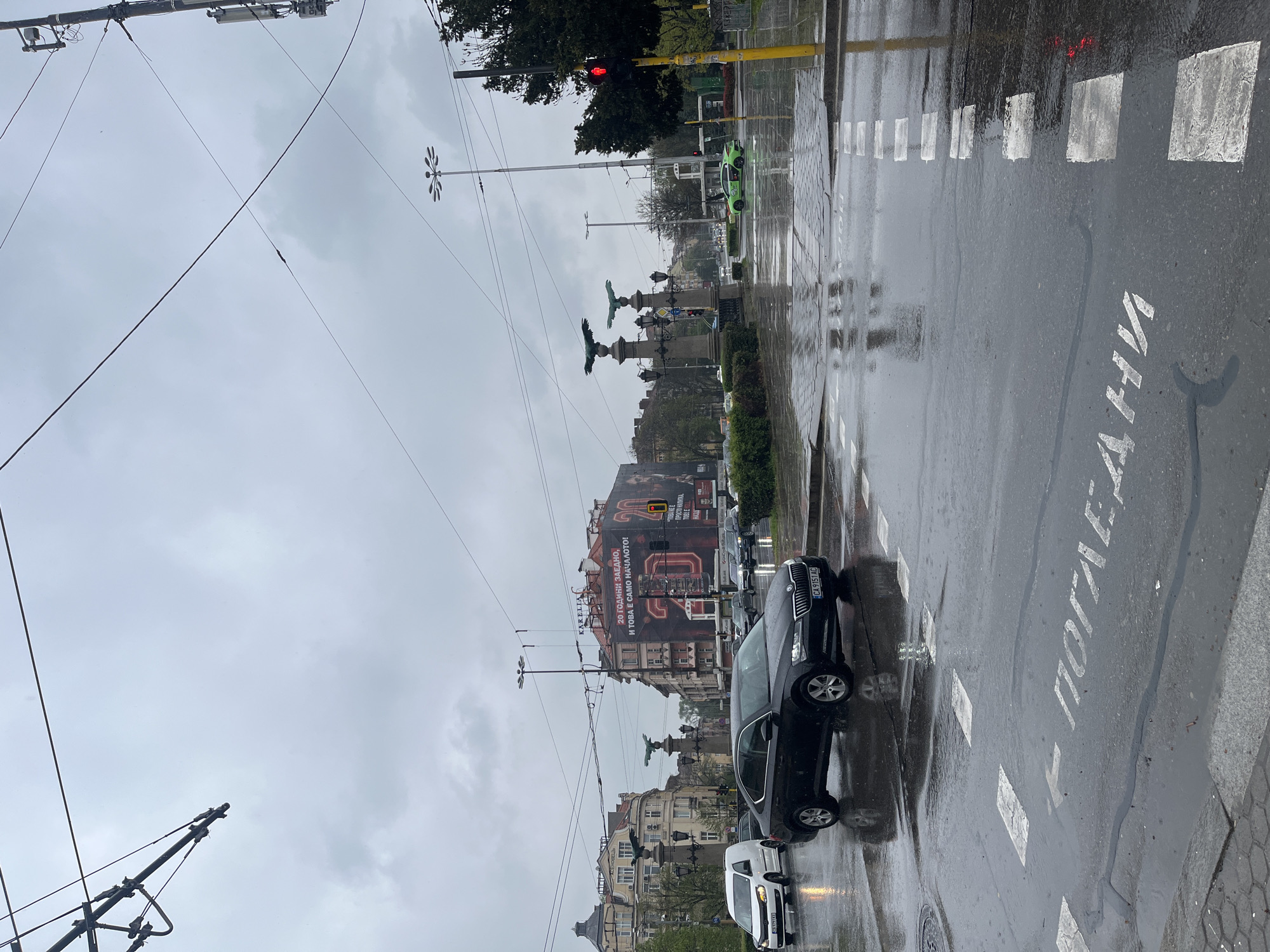 This media file captures a rainy, overcast day on a city street in Sofia, Bulgaria. The scene shows a wide street with wet asphalt reflecting the dull light, indicating recent rain.
In the foreground, a pedestrian crossing is marked with white rectangular stripes on the road. The Cyrillic word "ПОГЛЕДНИ" (POGLEDNI), meaning "LOOK" or "PAY ATTENTION," is painted in white on the road surface.
The midground features several vehicles, including a dark grey Skoda sedan with license plate "CA 9151 AC" and a white van, both positioned on the wet street. A bright green car is visible further down the road to the right. A red traffic light indicates that traffic is stopped. Overhead, a dense network of trolleybus or tram wires crisscrosses the sky, supported by numerous utility poles, some equipped with decorative, star-shaped light fixtures. A central median or island in the street is adorned with ornate lampposts and statues of winged figures.
The background consists of multi-story buildings with classic European architectural styles. One prominent building displays a large banner with Cyrillic text that reads: "20 ГОДИНИ ЗАЕДНО, И ТОВА Е САМО НАЧАЛОТО!" (20 YEARS TOGETHER, AND THIS IS JUST THE BEGINNING!), along with smaller promotional text: "ТОБА НЕ Е - ПРОСТО НАПИТКА, ТОБЛЕ" (THIS IS NOT - JUST A DRINK, TOBLE). The sky is a uniform, cloudy grey. No people are visible in the image.