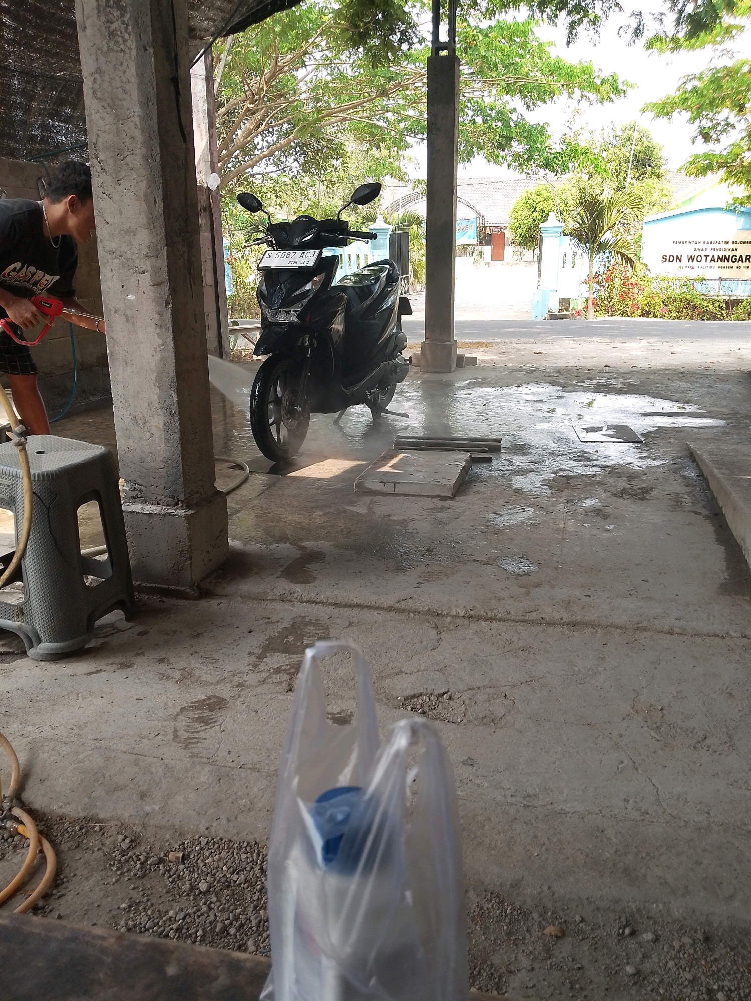 A young man wearing a black t-shirt and checkered shorts is pressure washing a black motorcycle in a covered outdoor area. He is holding a red pressure washer wand, with water spraying onto the motorcycle's wheel and the concrete ground. A grey plastic stool is to the left of the man, with a yellow hose coiled near it. The background shows a street with buildings and trees, and a sign for "SDN WOTANGGAR" school in Kalitidu, Indonesia. The ground is wet from the washing. A transparent plastic bag with a blue cap is in the foreground.
