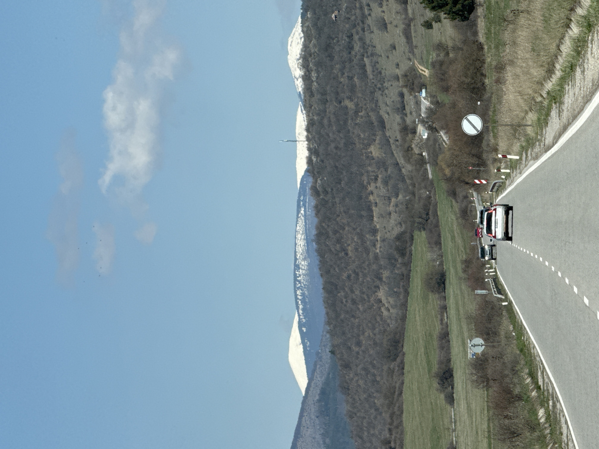 The image shows a road in Drenovo, Bulgaria, running alongside a mountainous landscape. The sky is blue with some clouds. Along the road, there is a white and red striped barrier, a road sign with a horizontal line through a circle, and a vehicle. The mountain is partially covered in snow. The roadside features a grassy area.