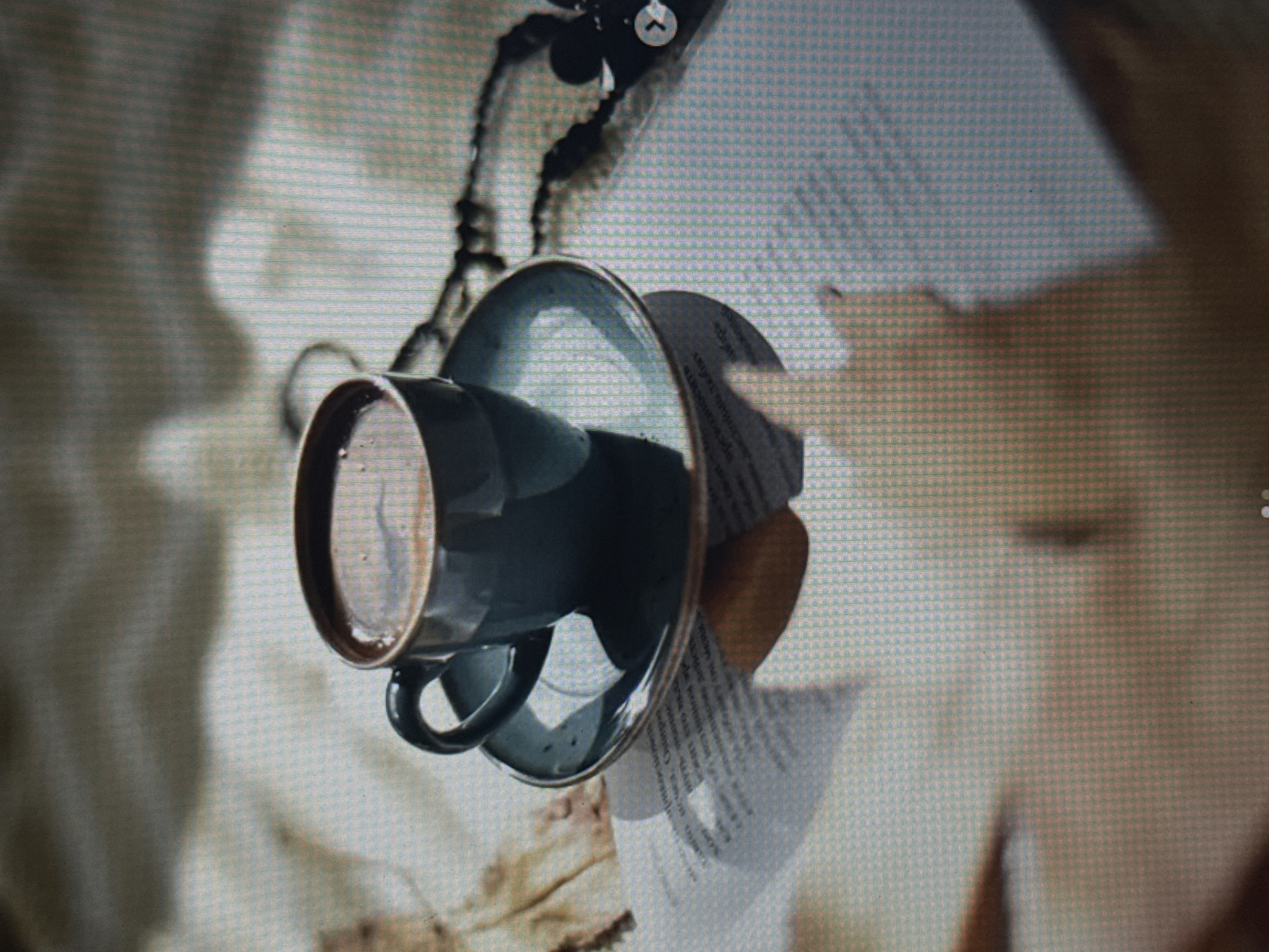 A close-up shot features a dark blue ceramic coffee cup and saucer resting on a surface that appears to be a page of text, possibly a book or magazine. The cup is positioned at an angle, with its contents, a dark liquid, visible. The saucer is beneath the cup, and a portion of its rim is in focus.
Behind the cup and saucer, there is a blurred background that suggests an indoor setting. The lighting appears to be soft, casting subtle shadows. The overall tone of the image is muted and artistic, with a focus on the textures and forms of the objects.
The text on the page beneath the cup is partially visible but not entirely legible. There are also some organic shapes, possibly dried leaves, scattered on the surface. A thin, dark, chain-like object is also visible, adding an element of intrigue. A small white circular icon with an upward-pointing arrow is also present in the upper portion of the image. No people are visible in the image. The context suggests a quiet moment of reflection or enjoyment of coffee, perhaps in a study or a cozy corner.