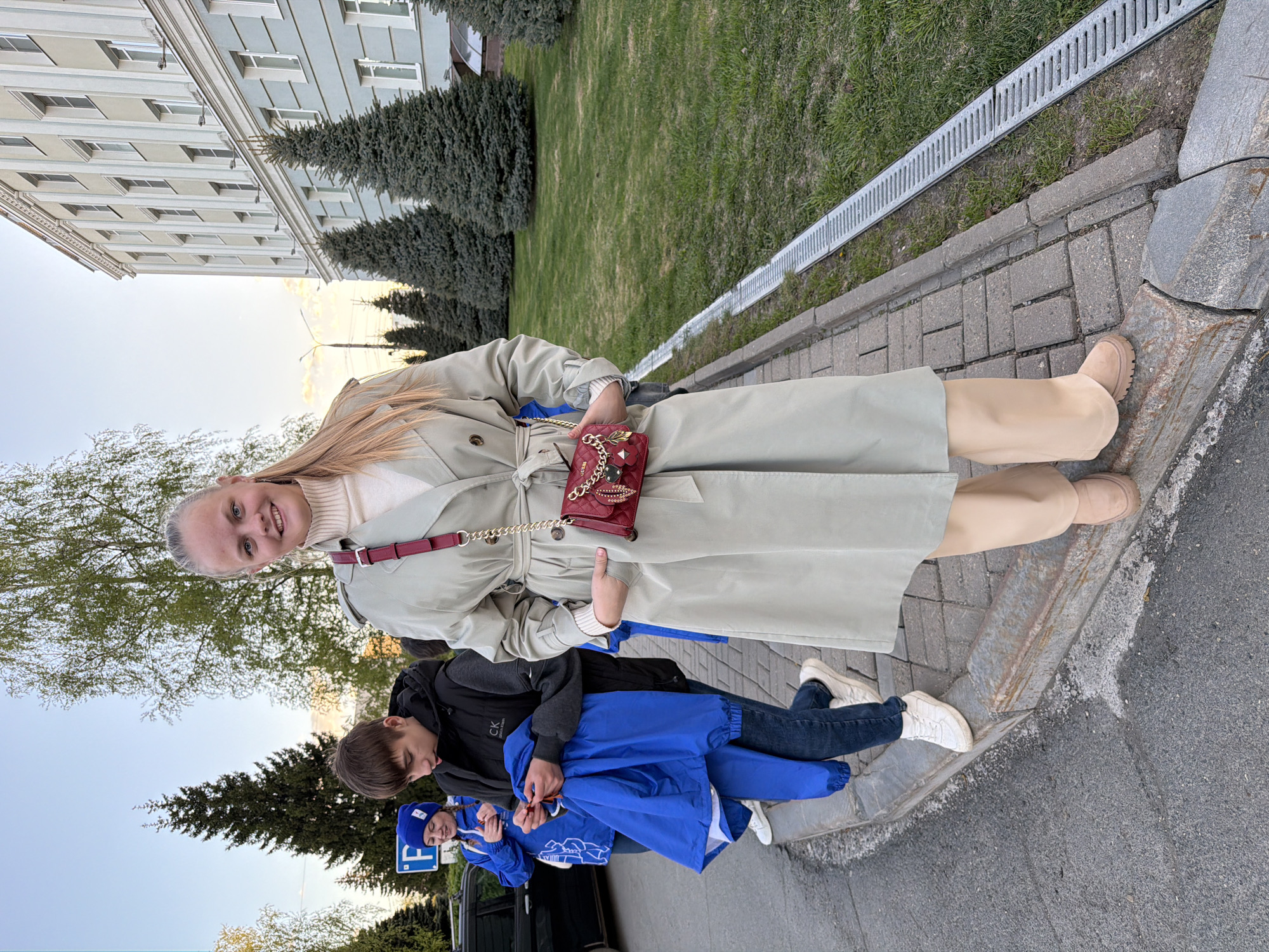 The image shows a woman in a light trench coat standing on a brick sidewalk in what appears to be a park or urban area. She is holding a red purse with a gold chain. Two young people are nearby; one is wrapped in a blue jacket. The environment includes green grass, trees, and a building in the background. The scene may be in Izhevsk, Russia.