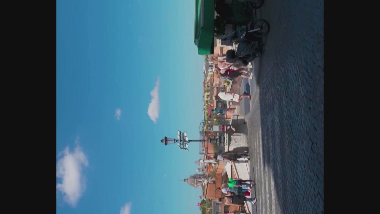 The image depicts a cobblestone street in an Italian city, with a slight upward slope towards a distant skyline featuring historical architecture and a prominent dome. The sky is a clear blue with scattered white clouds. In the foreground, a few people are walking on the street, including a group of women in casual attire and a man in a white shirt and shorts. To the right, a green-covered motorized vehicle, possibly a tourist cart or a food vendor's stall, is parked next to a scooter. A lamppost with multiple lights stands near the center of the frame. In the background, more people are visible walking on a stepped area leading up to buildings with terracotta roofs. The scene conveys a lively atmosphere typical of a tourist area. The provided crops do not contain specific information to identify the location as Morlupo, Italy.