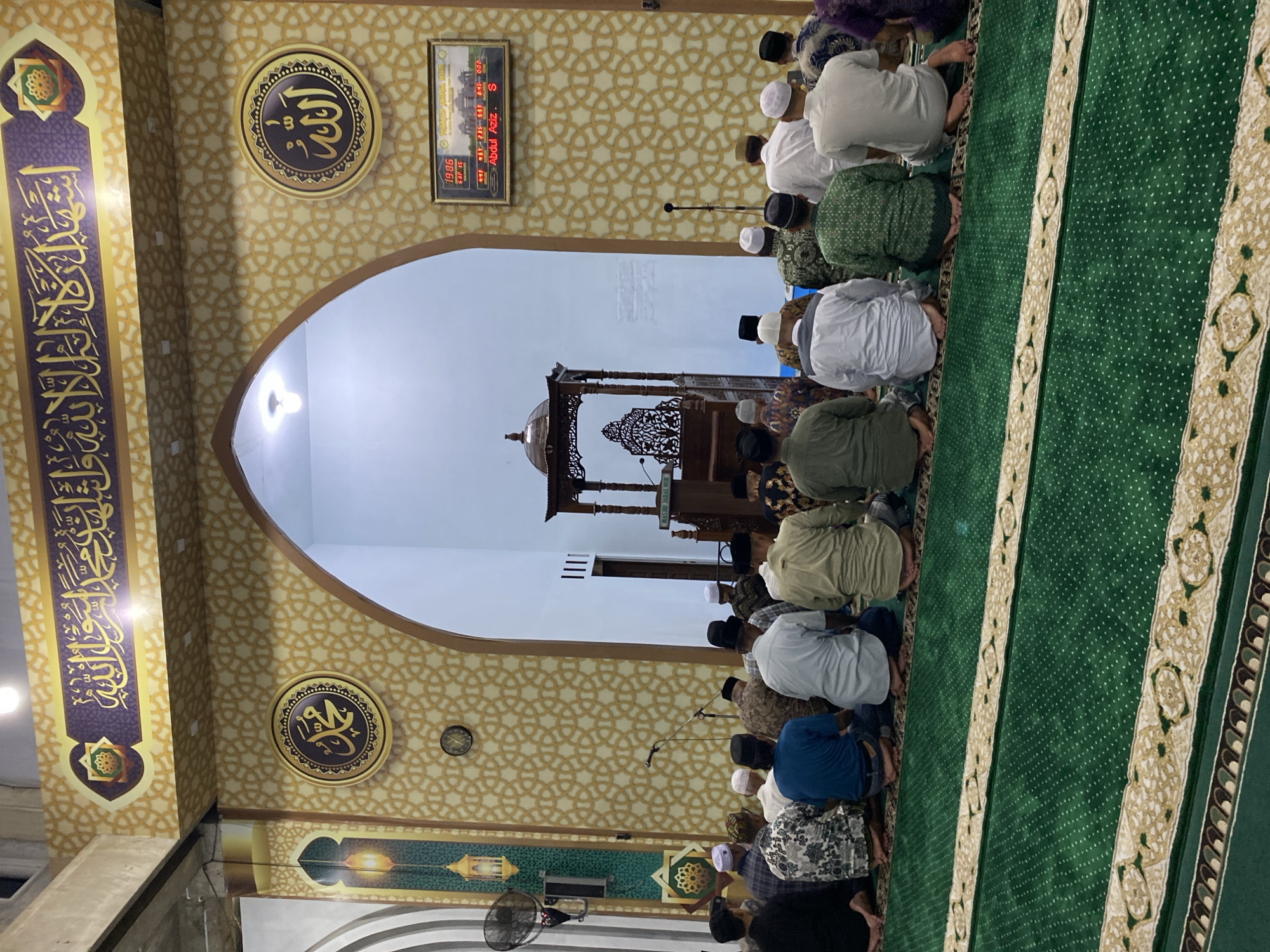 This is a medium shot, taken at eye level, inside a mosque. The mosque's interior is decorated with geometric patterns and Islamic calligraphy. In the foreground, a group of men are seated on a green carpet, facing away from the camera, towards an arched opening. They appear to be engaged in prayer. The men are dressed in traditional Islamic attire, including skullcaps and robes.
In the background, through the archway, a wooden minbar (pulpit) is visible. To the right of the archway, a digital clock displays the time "19:06". The mosque appears to be well-lit, with bright overhead lighting. The overall atmosphere is one of reverence and community.
This mosque is identified as Masjid Jabal Nur in the city of Lhokseumawe, Indonesia, based on the digital clock display.