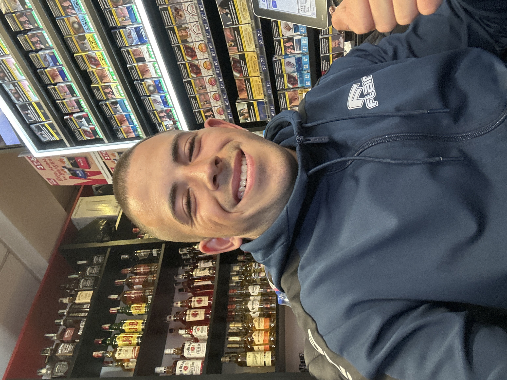 The image is a selfie of a smiling man with a shaved head, taken inside what appears to be a shop in Sofia, Bulgaria.
Behind the man, there are shelves stacked with cigarettes and liquor bottles. The cigarettes are packaged in boxes with various brand names and health warnings, some labeled 'Camel'. The alcohol bottles include brands like 'Jim Beam', 'PADEV', and 'КАРНОБАТ'.
The man is wearing a blue hooded sweatshirt with the word 'Flair' and a logo on the chest. Prices and labels are visible on the shelves behind him. The lighting is bright and the shelves are well-stocked.