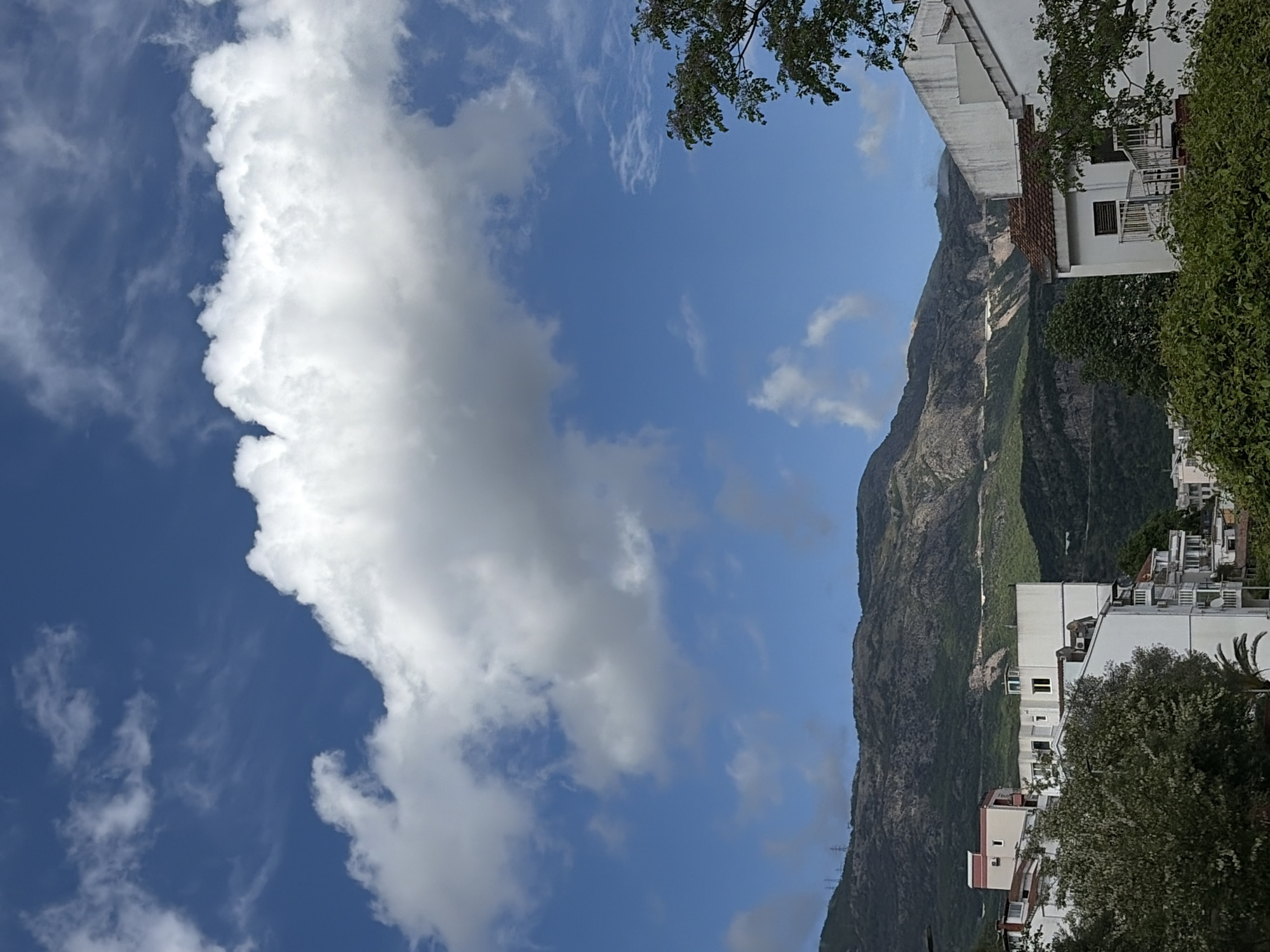 The image captures a bright, partly cloudy day in Budva, Montenegro. The sky is predominantly blue with scattered white and grey clouds dominating the left side of the frame. On the right, a rugged, steep mountain rises, covered with greenery and interspersed with rocky cliffs. At the base of the mountain are closely built white buildings with balconies, indicating residential areas. Trees and shrubs are interspersed among the buildings, adding patches of green. Tree branches are also visible in the upper-right corner. High-voltage power lines can be seen running alongside the mountain.