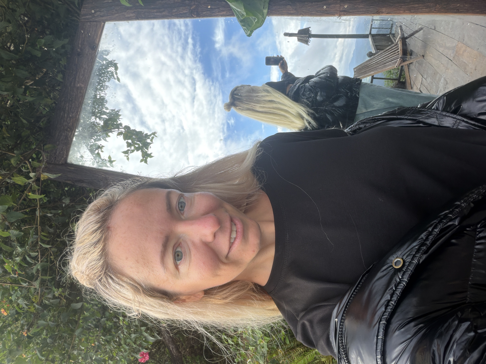 In Phát Chi, Vietnam, a blonde woman is taking a selfie in front of a large mirror. She is wearing a black shirt and a black puffer jacket. The mirror reflects the sky, clouds, and a mountain landscape. In the reflection, another person with blonde hair tied in a bun is visible, holding a smartphone. A wooden deck with a chair is also visible in the reflection. The woman's face is turned towards the camera, and she is smiling. The background behind the woman is filled with greenery.