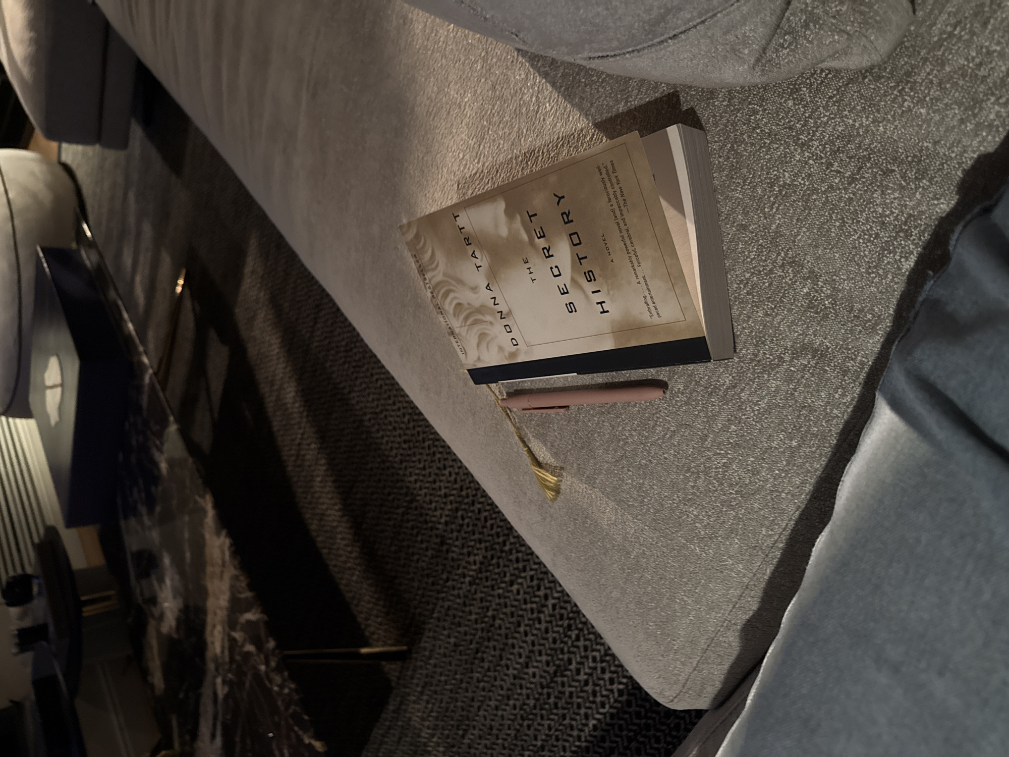 Here is a detailed description of the image:
The image shows a top-down, close-up view of a book titled "The Secret History" by Donna Tartt resting on a gray, textured couch. A pale pink pen and gold-colored tassel bookmark are lying on top of the book. The background is blurred, but a dark surface with a patterned texture can be seen. A portion of a person's leg clad in blue denim is visible on the right side of the frame. Based on the style of the furniture and the lighting, it is likely to be in a residential indoor setting. However, there is no additional information about the specific location in Mexico City, Mexico, or details about people, interactions, or actions.