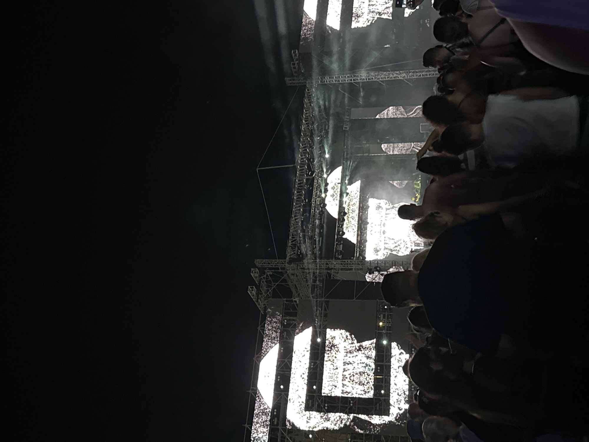 The image depicts a large crowd of people gathered at an outdoor night event, likely a concert or festival. The background features a prominent stage setup with large screens displaying abstract, pixelated imagery, resembling white or light gray silhouettes against a dark background. Several large metal scaffolding structures support the lighting and sound equipment. Beams of light and smoke are visible, suggesting a dynamic light show. The foreground is filled with the backs of numerous individuals, their faces mostly obscured by darkness and their positioning in the crowd. They appear to be engaged in watching the stage, with some individuals in the mid-ground visible from the side or front, suggesting they are dancing or interacting. The overall atmosphere is dark, illuminated primarily by the stage lights and screens. The location is described as Unknown, Unknown.