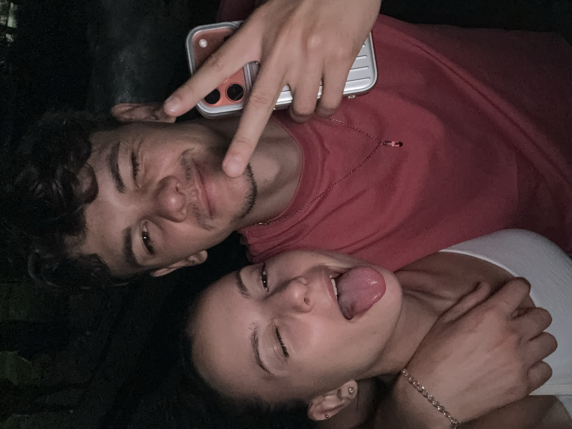This is a selfie of a young couple embracing outdoors at night, likely in Sandanski, Bulgaria. The woman on the left is winking, with her tongue sticking out playfully. She has dark hair pulled back, a bracelet on her left wrist, and small earrings. The man on the right is also winking and holding a smartphone in his right hand, with his fingers forming a peace sign. He has curly dark hair and is wearing a red t-shirt with a silver necklace. The background is dark, suggesting it is nighttime, possibly in a park or wooded area.