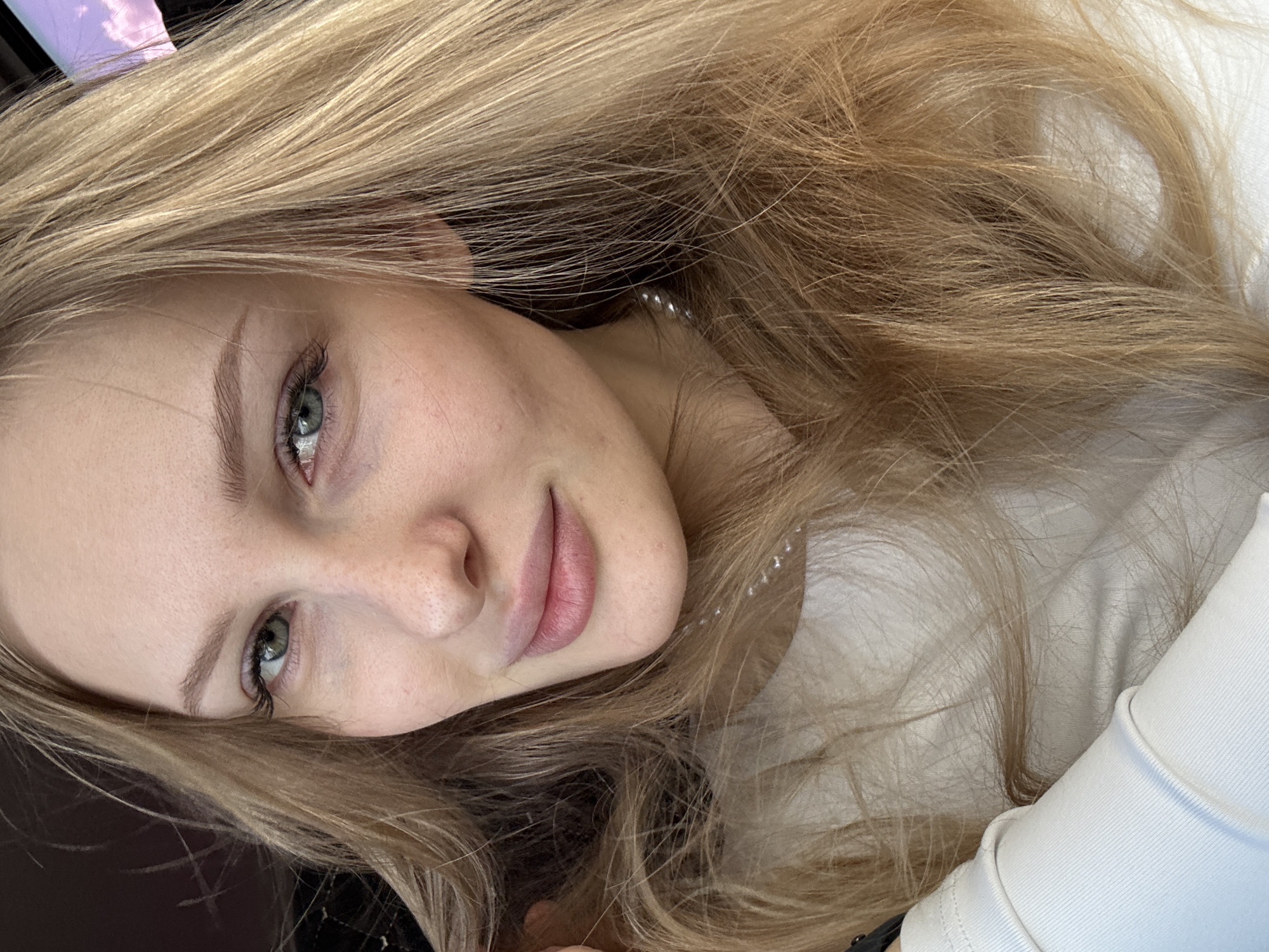 A close-up portrait features a young woman with fair skin and long, wavy, golden blonde hair that largely frames her face and fills the background. She has light-colored eyes, possibly blue or green, with prominent eyelashes and well-groomed eyebrows. Freckles are visible across her nose and cheeks. Her lips are a natural pink color, slightly parted in a gentle smile. She looks directly at the camera with a calm and serene expression.
She is wearing a white, long-sleeved top or sweater, visible on her right side, and a delicate necklace composed of small, round pearls or beads is subtly visible around her neck. Her hair spreads around her head, with some strands covering parts of her clothing and the background. Hints of the background suggest an indoor setting or a vehicle, with a dark surface on one side and a lighter, possibly white, surface on the other. A subtle purple object is partially visible near the top left of the frame. The lighting is soft and diffused, highlighting her features without harsh shadows. No text is visible in the image. The person is located in Perm, Russia.