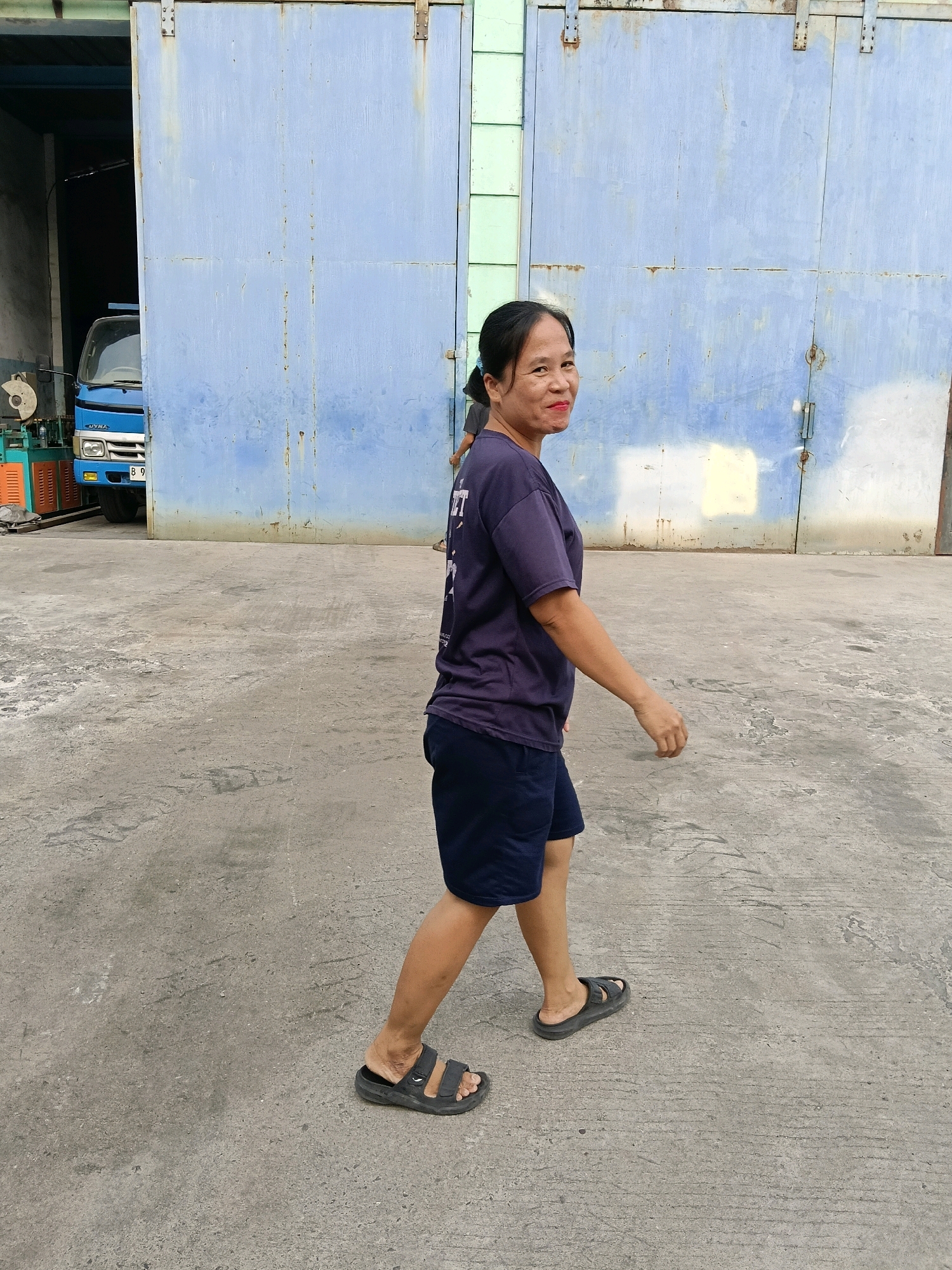 A woman with black hair tied in a ponytail, wearing a dark purple t-shirt and navy blue shorts, walks across a concrete surface. She is wearing black sandals. She turns her head to look back over her right shoulder with a slight smile. Behind her, a large, pale blue sliding door, appearing to be made of metal, takes up a significant portion of the background. The door shows signs of rust and wear. To the left of the sliding door, a blue truck is partially visible, parked inside what appears to be a building or covered area. The environment suggests an outdoor industrial or storage area. The image does not provide specific location details for Belimbing, Indonesia.