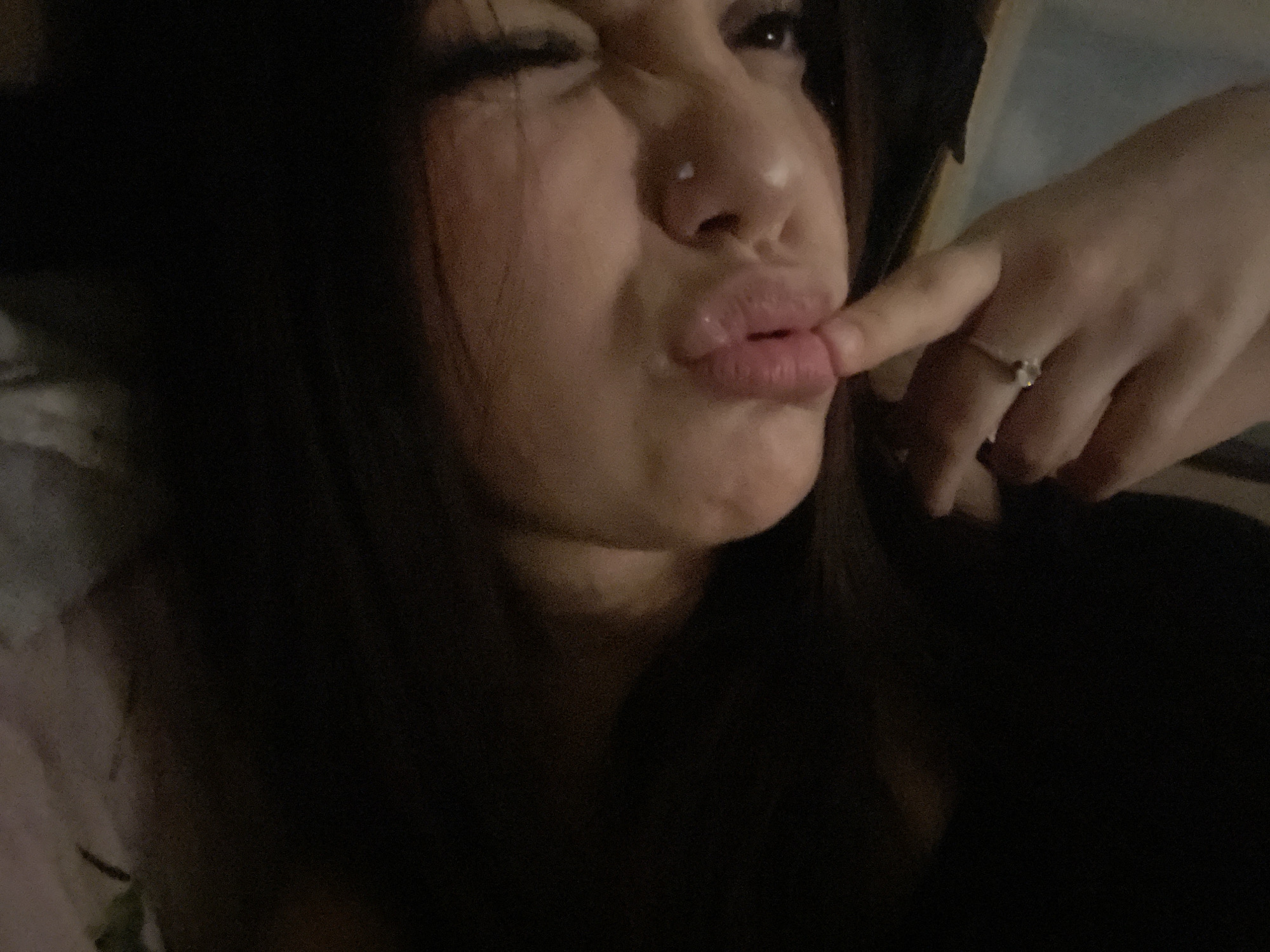 A close-up, low-light photograph captures a person, likely female, making a playful or exaggerated face. The subject's dark, long hair frames her face, which is positioned on the left side of the frame. Her left eye is squinted shut, while her right eye is partially open. Her lips are slightly puckered, and a finger is gently pressing against the corner of her mouth. A small nose stud is visible on her right nostril. The finger belongs to a hand (possibly her own or another person's) visible on the right side of the image, wearing a ring with a small, clear stone on the ring finger. The lighting is dim, suggesting a nighttime setting or a dark indoor environment. The background is dark and largely out of focus, providing minimal contextual details beyond the immediate subjects. No text is visible. The location context for this media file is Saint Petersburg, Russia.