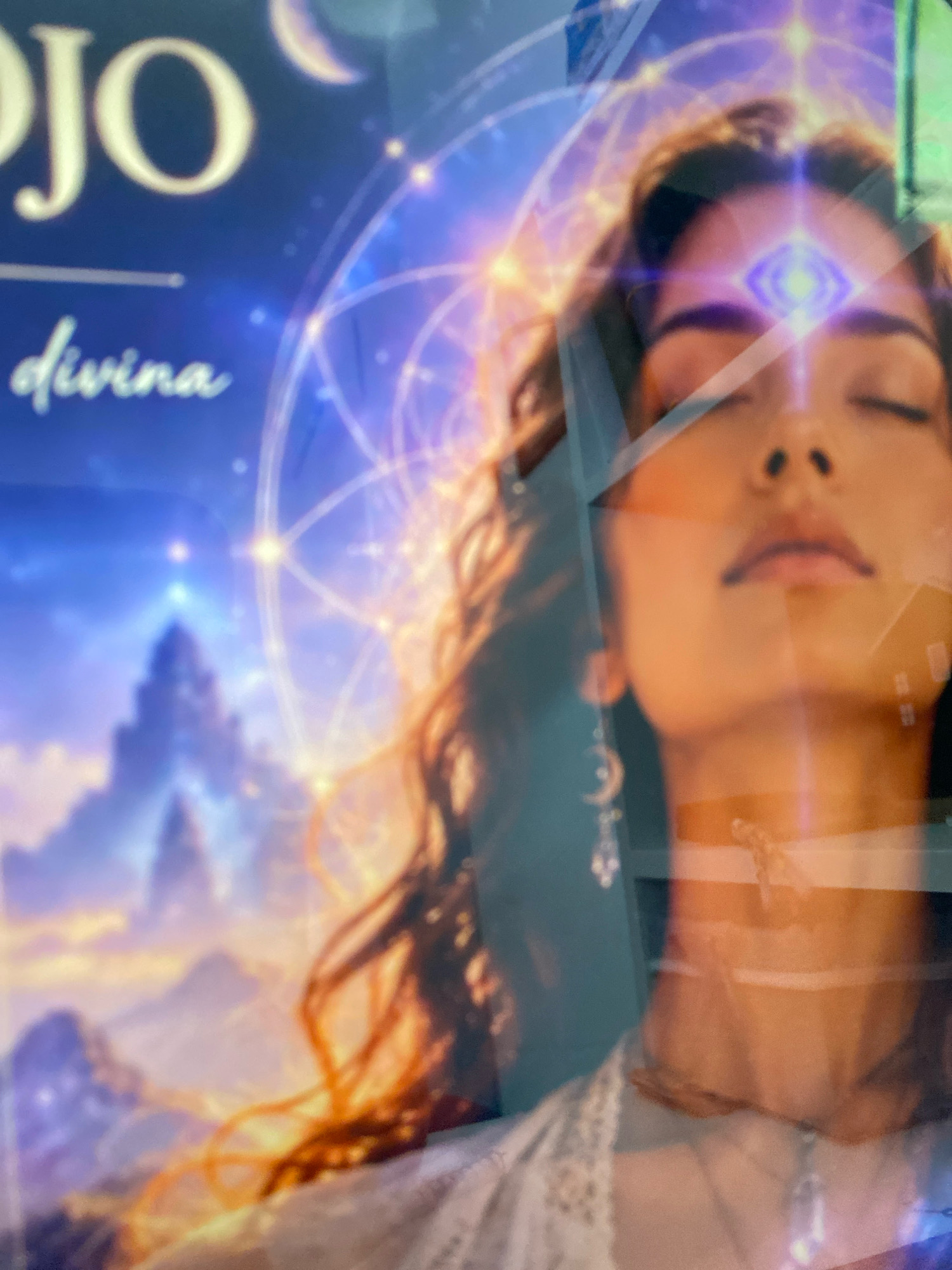 The image is a close-up shot of a poster or artwork depicting a woman's face, overlaid with spiritual and cosmic imagery. The woman's eyes are closed, and she has a bright, glowing symbol on her forehead, possibly representing a third eye or spiritual energy. She wears delicate jewelry, including earrings with crescent moon charms and a necklace. Behind her, there are intricate geometric patterns and celestial elements, suggesting a theme of spirituality, mysticism, or astrology. In the background, there's a blurry depiction of a mountainous landscape with a castle-like structure. The text "JO" and "divina" are visible on the left side of the poster in a stylized font. The lighting is warm and ethereal, with purples, blues, and golds dominating the color palette. The overall impression is one of serenity and cosmic connection. The image is not set in a specific location in Colmenar, Spain, but appears to be a piece of art displayed on a wall or screen, potentially in a commercial or public space.