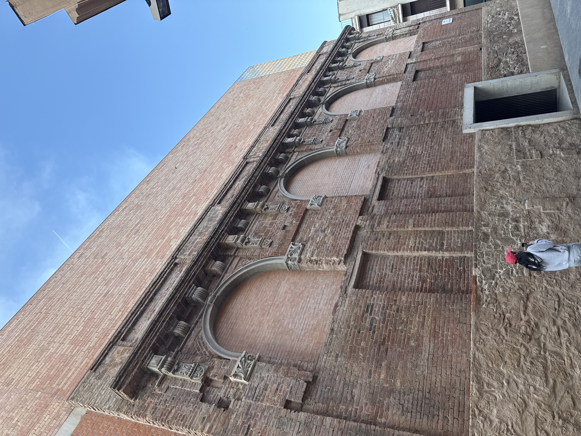 A low-angle shot reveals a tall, brick building facade in Barcelona, Spain, under a clear, blue sky. The structure features three prominent archways, seemingly filled in with brick, stacked vertically along one side. The brickwork varies in color and texture, suggesting possible repairs or additions over time. A section of the wall adjacent to the archways is constructed of a rougher, stone-like material. A rectangular opening or window is visible near this stone section. In the corner of the image, a person wearing a grey sweatshirt and a bright red hat is visible; the person is holding a phone, and is likely taking a picture of the building. The overall architectural style is classical with ornate details around the arches. Other buildings are partially visible at the top of the frame.