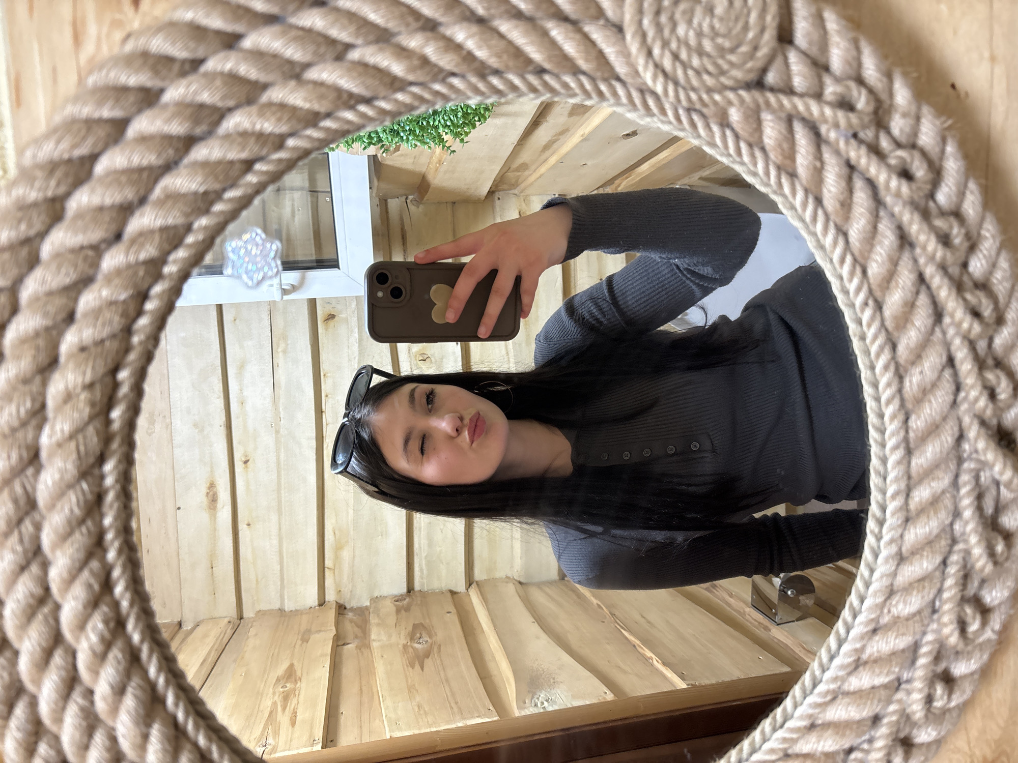 A person with long dark hair, wearing a gray, buttoned top and sunglasses atop their head, is taking a selfie in a rope-framed mirror. They are puckering their lips and winking. They are holding a brown phone case with a light brown heart on it. The surrounding environment appears to be a room with light wood paneled walls. A window with a white frame and decorative light-catching ornament and some greenery outside is visible. The exact location of this image is unknown.