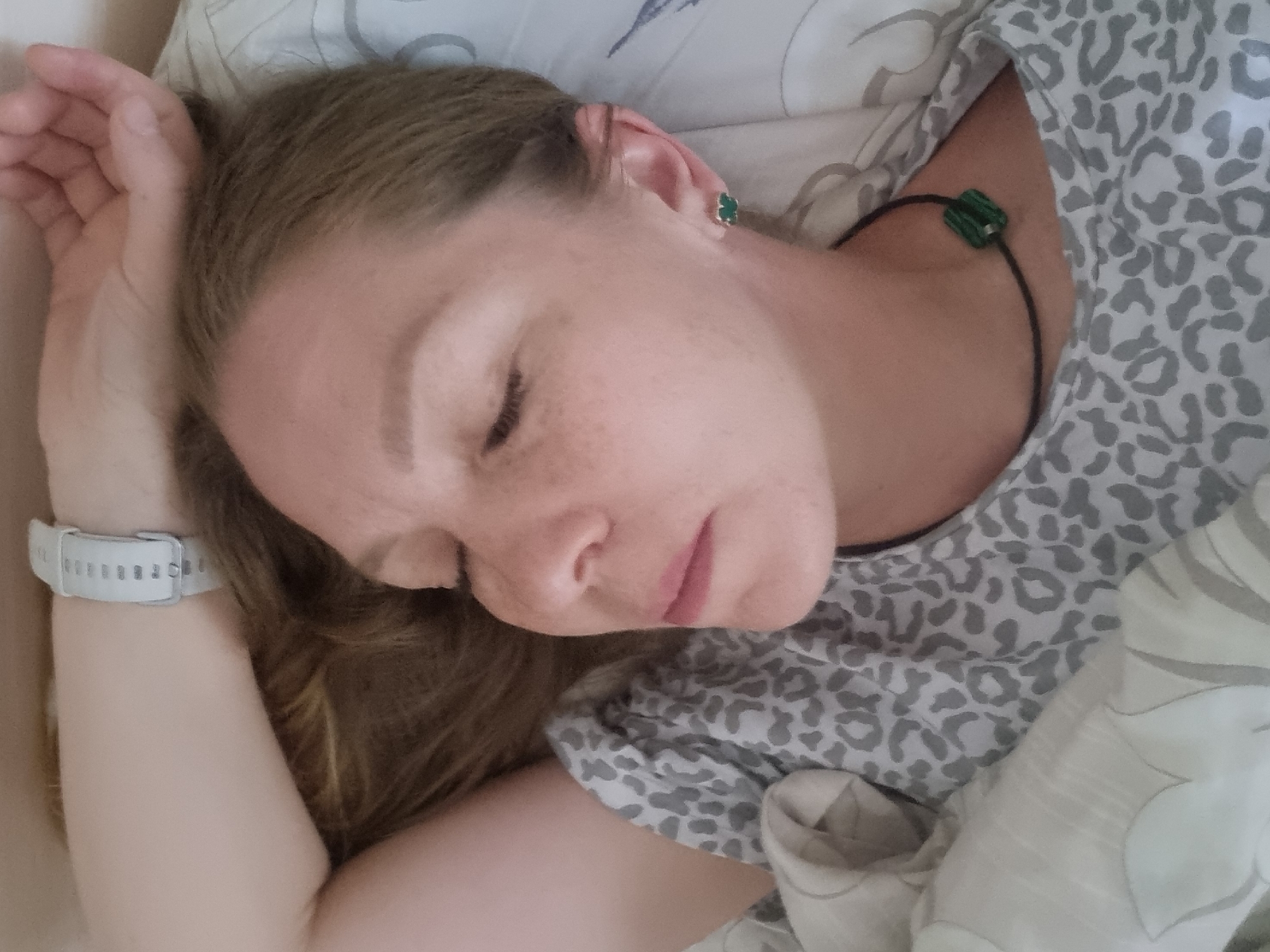 A woman with fair skin and light brown hair is sleeping in a bed. She is lying on her side, facing left. Her left arm is bent and resting near her head. She is wearing a white smartwatch on her left wrist and a green stone earring in her right ear. Around her neck, she wears a black cord necklace with a square green pendant. She is wearing a gray and white animal print top. The bed has a light-colored pillow and patterned bedding. The location is unknown.