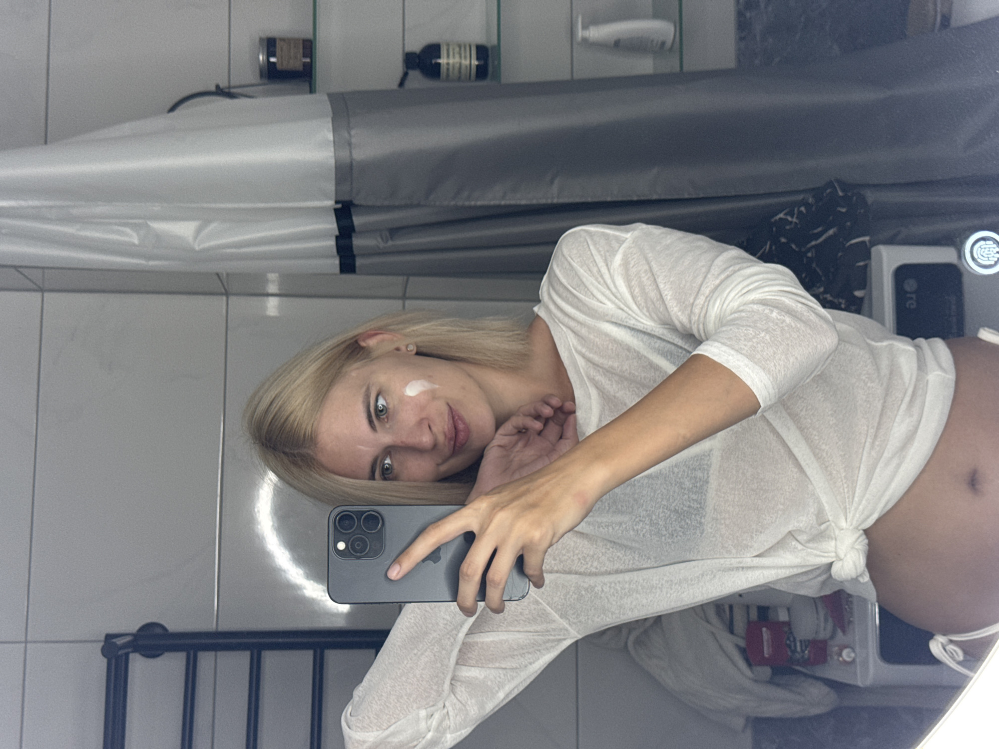 A blonde woman is taking a mirror selfie in a bathroom. She is wearing a sheer white long-sleeved shirt tied at the waist, revealing her abdomen. There is a patch of white cream on her right cheek. She is holding a smartphone in her left hand, which is positioned near her face. The bathroom has tiled walls, a shower curtain, shelves with various bottles and jars, and a towel rack. The image does not contain any information about the city of Saratov, Russia.