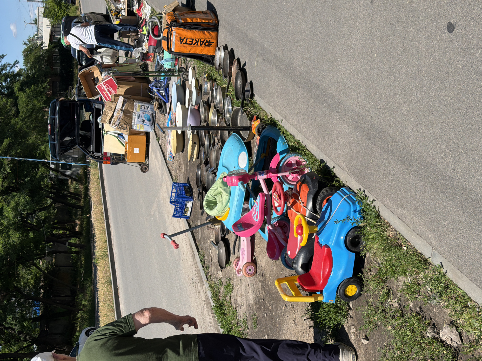 A street scene in Dnipro, Ukraine, shows items laid out for sale along the side of a road. In the foreground, a person in a green t-shirt and dark pants stands on the grassy shoulder, facing away from the camera. To the right of the person, various toys are arranged on the ground, including a blue and red ride-on car, a pink tricycle, and a blue child's seat. Behind the toys, a collection of metal pots and pans are stacked and spread out. Further back, several car trunks are open, revealing more items for sale, including boxes, bags, and household goods. A person in a white shirt and green cap is visible near the open trunk of a car on the right, appearing to be engaged in the sale. The environment is urban, with a paved road, sidewalk, and trees lining the street. The sky is clear and blue.