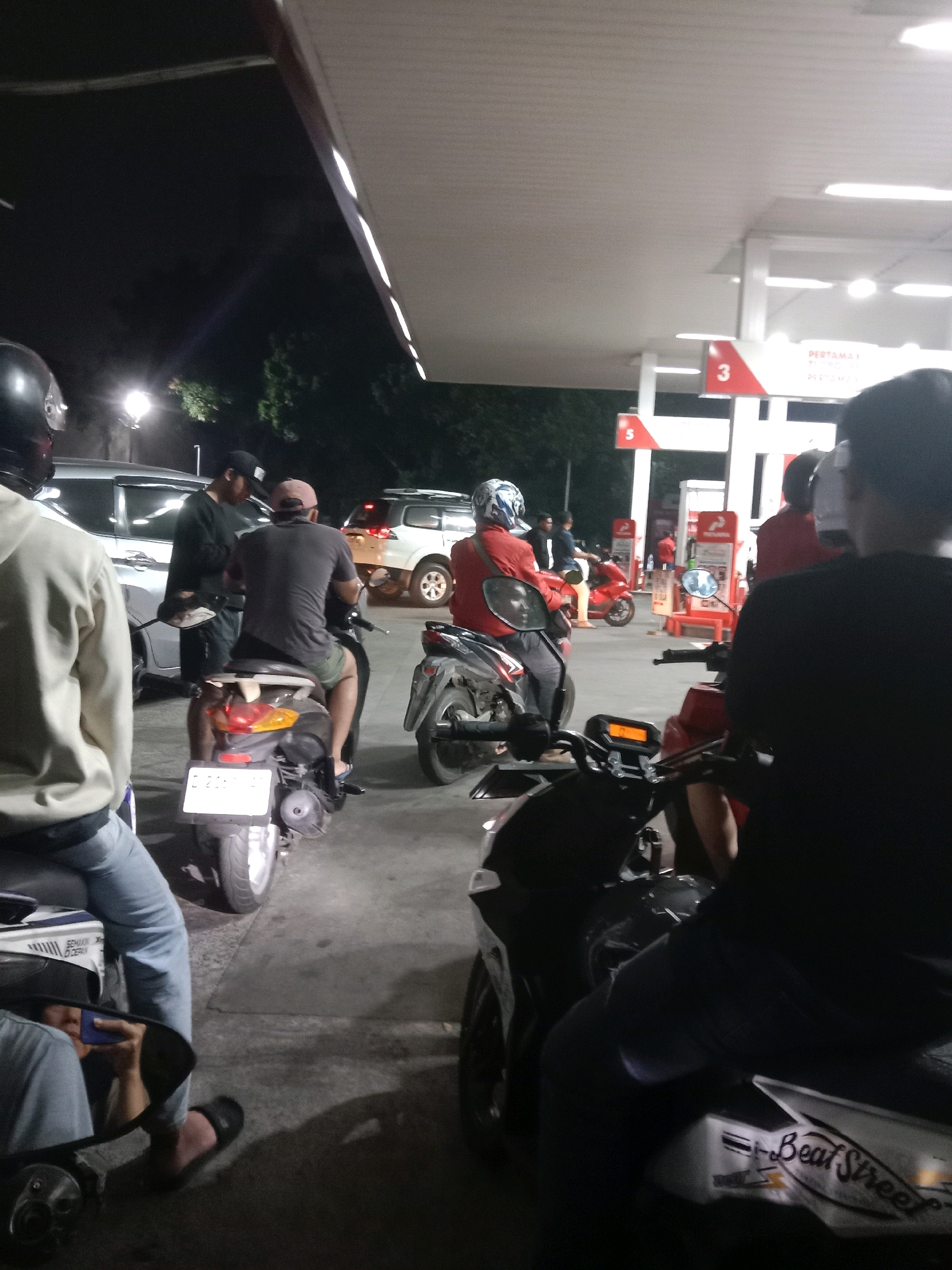 The image depicts a nighttime scene at a Pertamina gas station in South Jakarta, Indonesia, with several motorcycles and a few cars present. In the foreground, a person in a black shirt is seated on a motorcycle, facing away from the camera, with another motorcycle to their left. Further back, a person in a light-colored hoodie and blue jeans is also on a motorcycle, partially obscured. Adjacent to them, another person is standing near a silver car, and to their right, a person in a gray shirt and a cap is seated on a dark-colored motorcycle. In the background, a white SUV is visible, and a red motorcycle is parked. The gas station has multiple pumps, with signage indicating "PERTAMAX". The overall atmosphere suggests a typical evening at a busy roadside establishment.