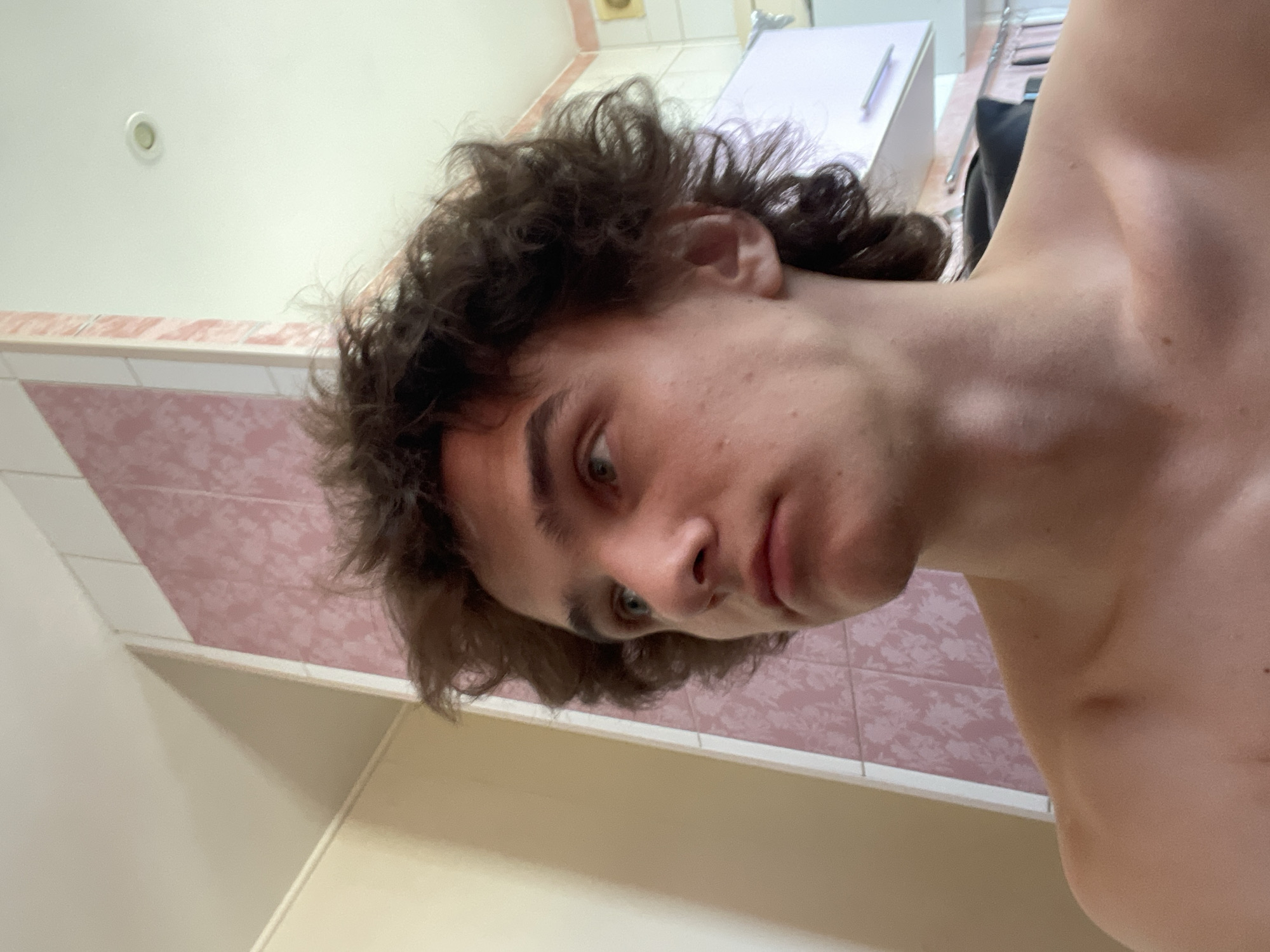 A close-up, slightly skewed angle photograph shows a light-skinned person with curly, dark brown hair. The person's face is angled toward the camera, revealing light-colored eyes, a nose, and closed lips. The person is shirtless, with a portion of their shoulder visible in the right of the frame.
The background includes a wall partially covered in tiles with a pink floral pattern. A portion of a ceiling with a recessed light fixture is visible in the upper left. Other indistinct objects, including what appears to be a white cabinet or counter, are located in the blurred background.
The location is likely a bathroom or washroom. The image does not have enough data to confirm the location within the city of Krasnoyarsk, Russia.