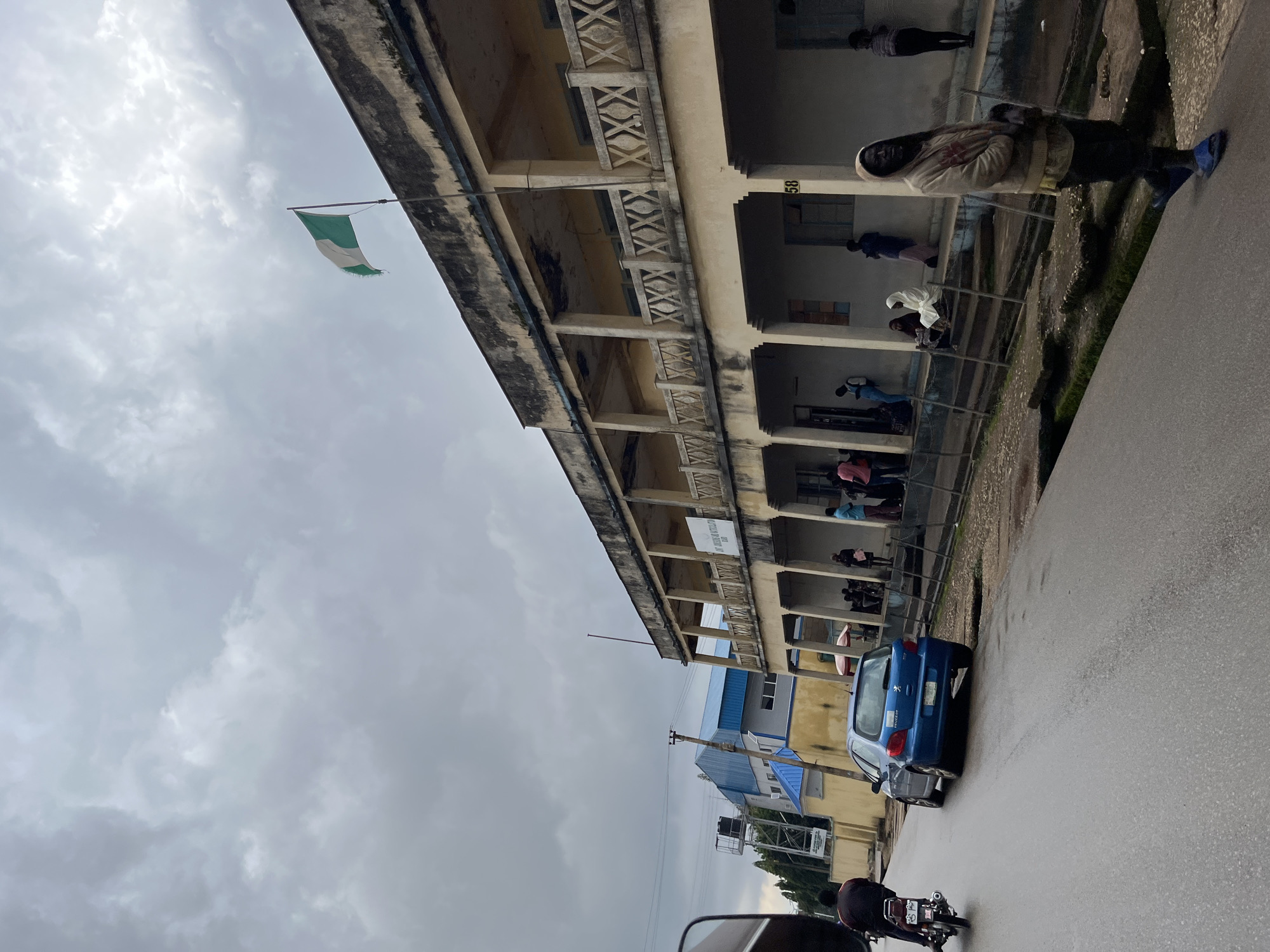 The image captures a street scene in Jos, Nigeria, under a cloudy sky. On the right, a long, two-story building with a weathered facade and a covered walkway supported by pillars dominates the view. The upper level has a decorative railing with an "X" pattern, and the lower level has open entrances. A Nigerian flag is visible flying from a flagpole atop the building. Several people are gathered outside the building, some sitting on steps, others standing or walking. On the left side of the street, a blue Peugeot car is parked. A person on a motorcycle is traveling away from the camera. Further down the street, another building with a blue, sloped roof is visible. The road is wet, suggesting recent rain. The overall impression is a typical urban environment in Jos on a somber day.