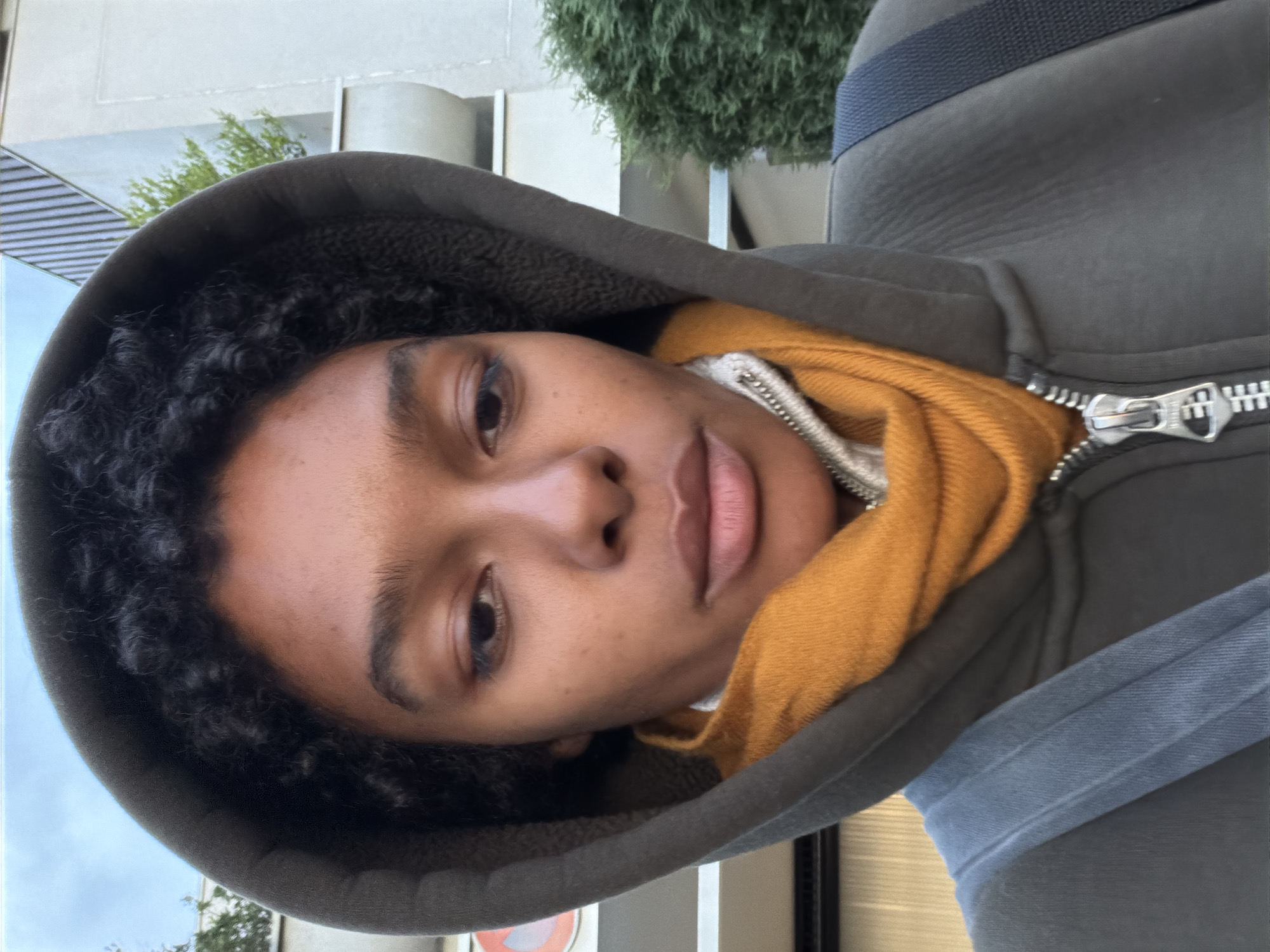 A close-up, rotated image shows a person with brown skin and curly black hair, wearing a gray hooded sweatshirt over a yellow scarf. The zipper of the sweatshirt is partially visible. A blue strap is seen across the person's shoulder. Behind the person, out of focus, are a building and green foliage. There is no indication of the city of Champs-sur-Marne in the image.