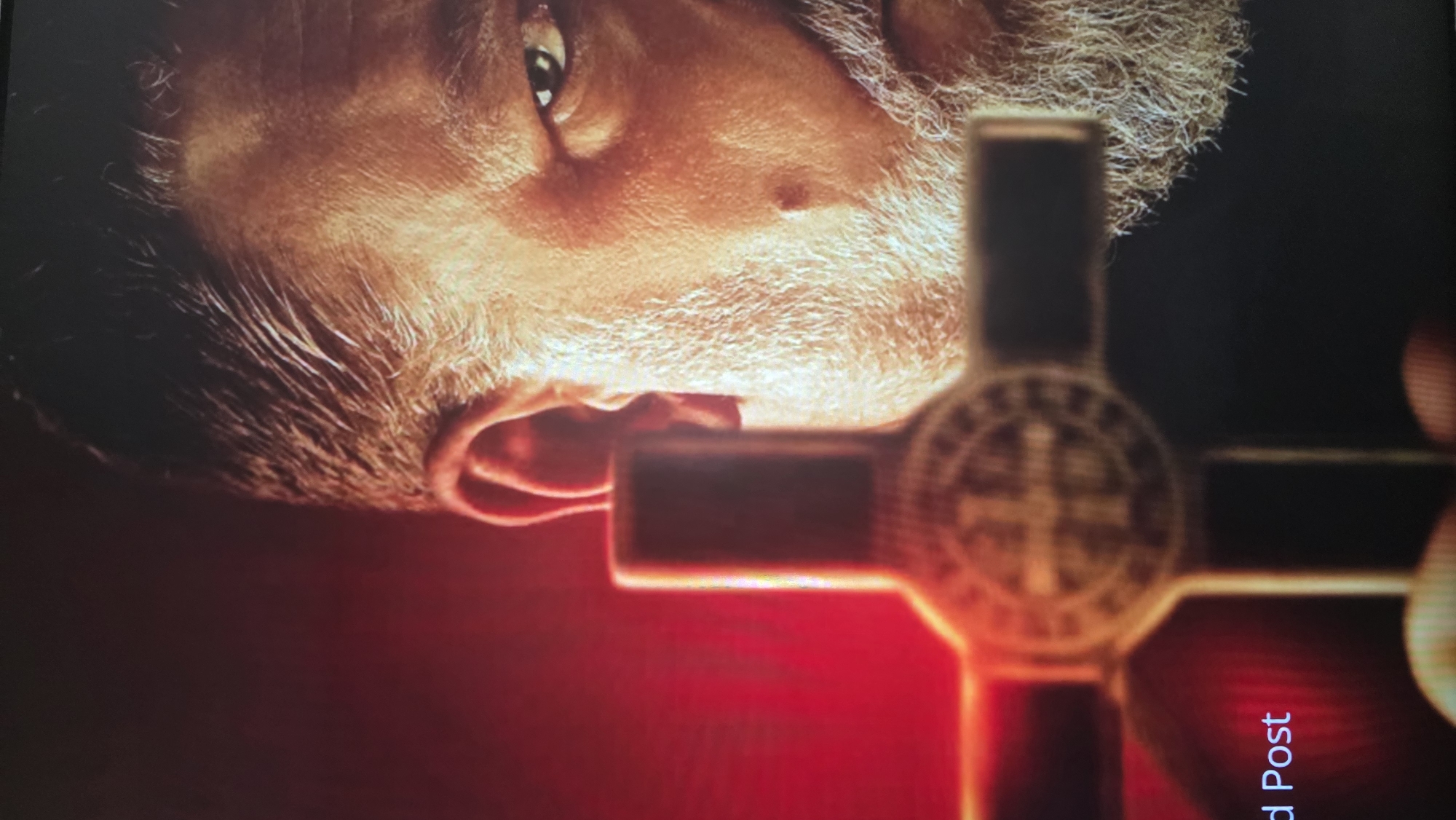 The image shows a close-up of a man's face next to a blurred Christian cross. The man has a light complexion, with visible facial hair including a white beard and mustache. One eye is visible, and there's a mole near his nose. The lighting is dramatic, with red hues dominating the background and parts of the man's face. The cross is brass colored with a prominent circular design in the center. No context indicates a location in Blagoevgrad, Bulgaria.