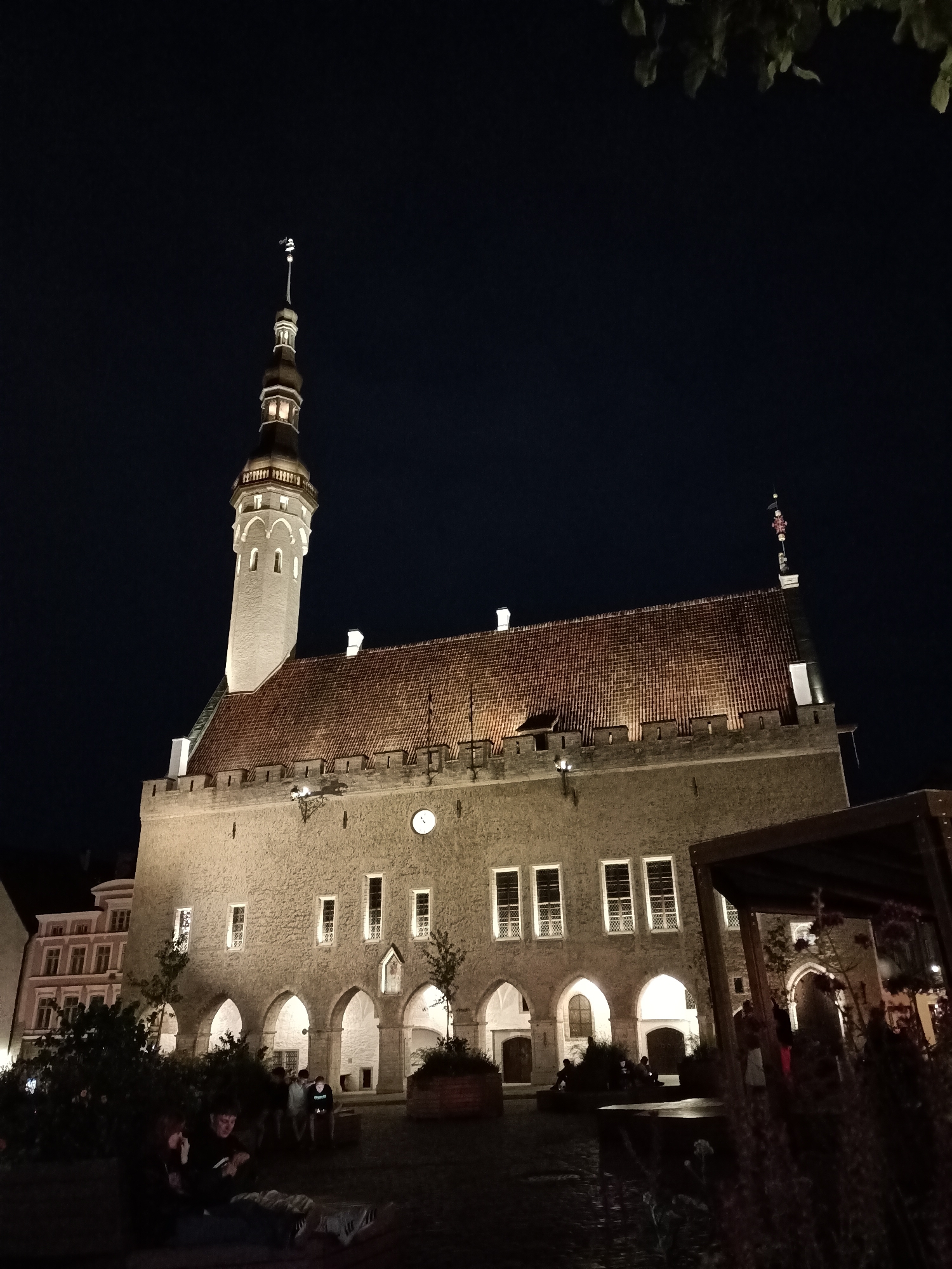 The image shows the Tallinn Town Hall at night, illuminated by artificial lights. The gothic architecture of the building is visible, with its prominent tower and tiled roof. Several people are gathered in the square in front of the Town Hall, some sitting and others standing, interacting with each other. The square appears to be paved and there are some trees and benches. The sky is dark, indicating it is nighttime. The lighting on the building highlights its texture and architectural features, creating a dramatic effect. The scene suggests a social gathering or a leisurely evening in the heart of the city.