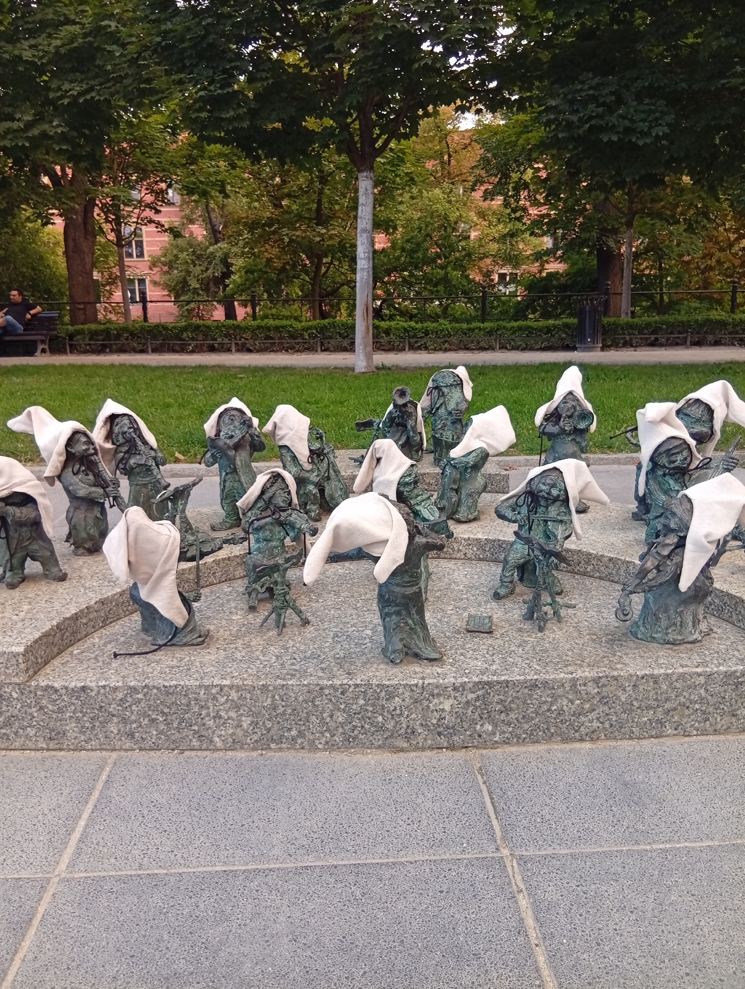 The image depicts a gathering of numerous bronze dwarf figurines, each wearing a white cloth draped over its head, positioned on and around a tiered granite structure. Some of the figurines appear to be holding miniature musical instruments, such as violins and trumpets. The setting is an outdoor public space, likely a park, with a grassy area and trees visible in the background. A red-brick building with windows can be seen through the foliage. In the far left background, a person is seated on a dark-colored bench, facing away from the camera. The figurines are arranged in a somewhat scattered but clustered manner, suggesting a communal scene. The ground in the foreground is paved with large, grey square tiles. The overall lighting suggests it is daytime, possibly late afternoon or early evening, given the soft light and shadows. This particular installation is a well-known feature of the city of Wrocław, Poland, specifically related to the "Wrocław Dwarves" (Wrocławskie Krasnale) which are a popular tourist attraction, often found in various locations across the city.