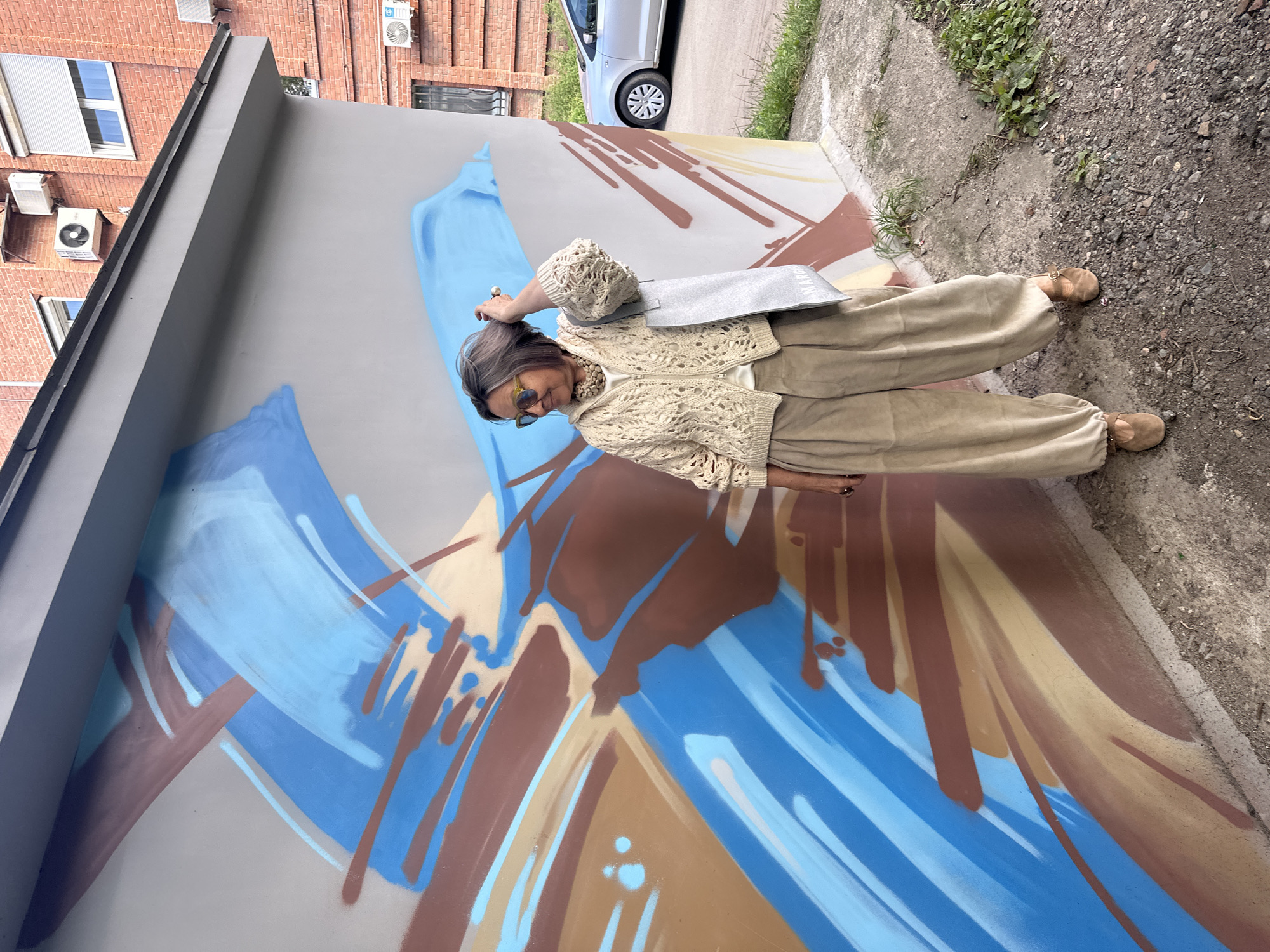 A medium shot captures a woman standing to the left of a brightly painted abstract mural on the side of a building. She is facing right, looking away from the camera with her right hand touching her hair. She wears a cream-colored knitted cardigan over a white shirt, loose tan pants, and brown slip-on shoes. A silver tote bag hangs from her right shoulder. The mural features bold strokes of blue, brown, and tan against a gray background. The building is constructed of red brick. To the right of the building, a silver car is parked on a concrete surface littered with gravel and vegetation. The scene appears to be an outdoor urban environment.
