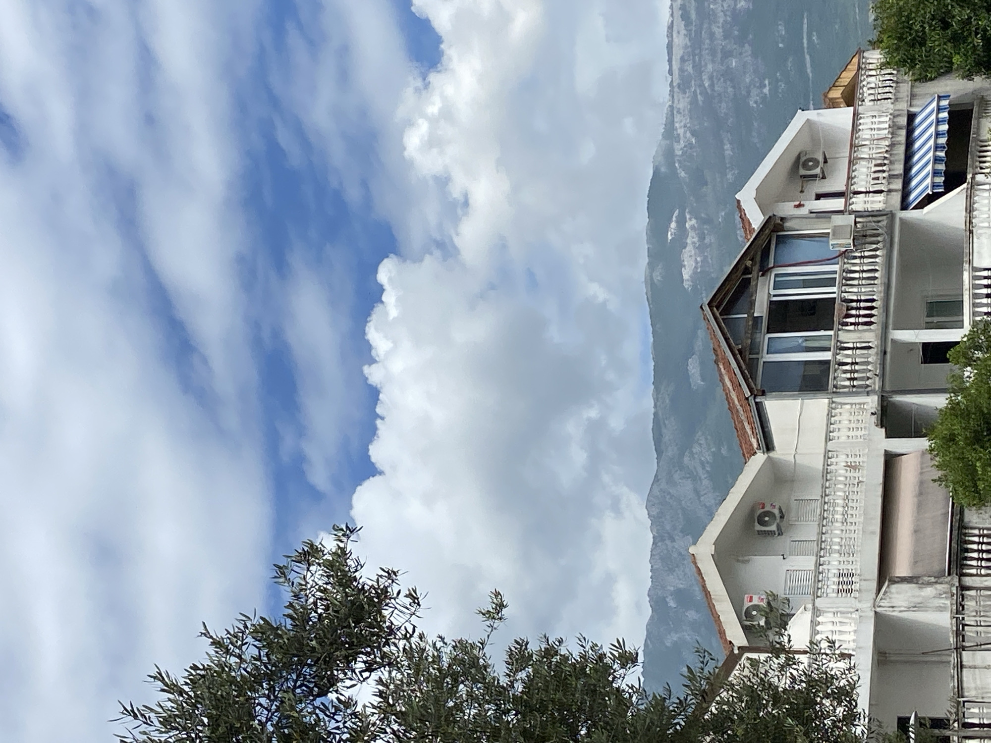 The image depicts a view in Budva, Montenegro. A multi-story white building with balconies is visible on the right side of the frame. The building has air conditioning units on its exterior walls. Behind the building, a mountain rises. The left side of the image is dominated by a blue sky with white clouds and the tops of some trees.