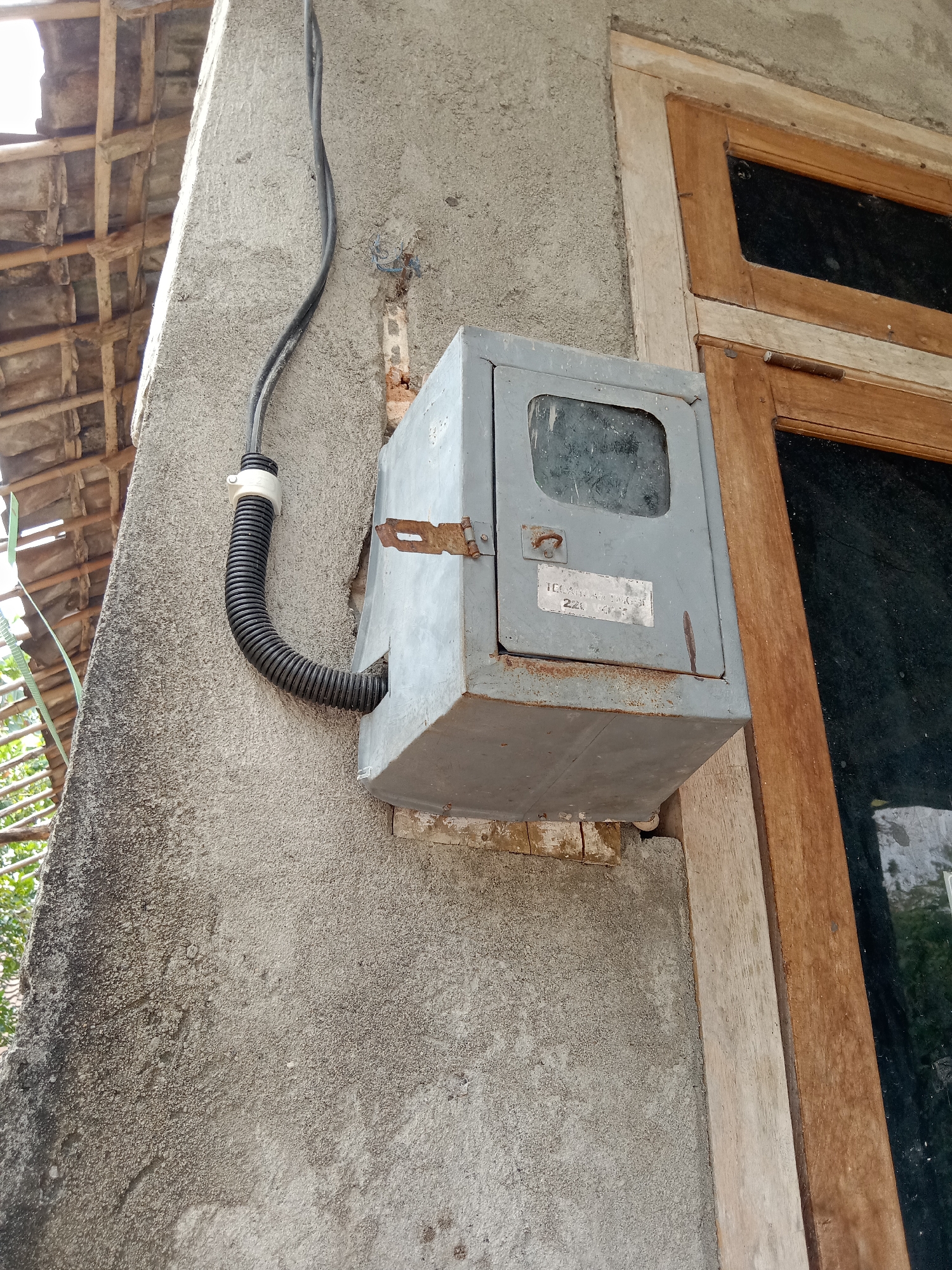 This is a close-up, eye-level, outdoor, daytime, medium shot of an outdoor electrical meter box. The meter box is gray, rectangular, and mounted on a stucco wall. A black, flexible conduit pipe connects to the bottom of the meter box and runs up the wall to an electrical cable. The wall is rough and textured, with some visible damage and discoloration. To the right of the meter box, a wooden window frame with a dark pane of glass is visible. Above and to the left of the meter box, a portion of a tiled roof supported by wooden beams can be seen. The overall scene suggests a residential or utility structure in an outdoor setting. The image was taken in the city of Jumput, Indonesia.