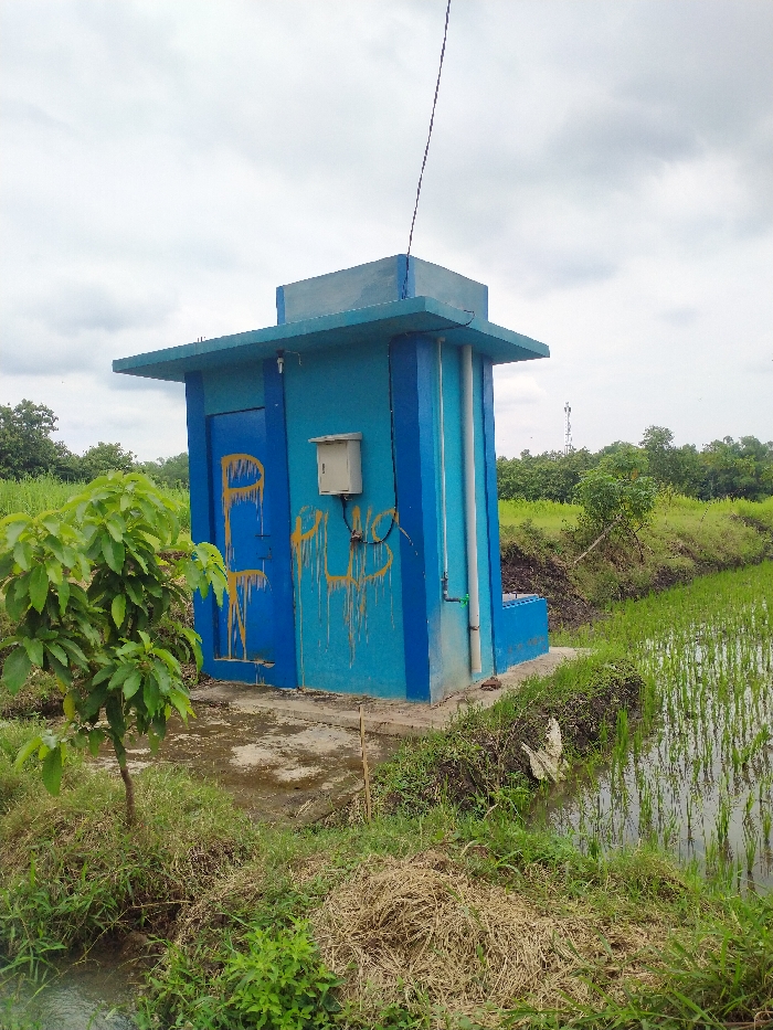 This image captures a small, utilitarian blue building situated amidst an agricultural landscape, specifically rice paddies, in Kecamatan Kebakkramat, Indonesia. The building itself is a bright aqua blue with darker blue trim on its edges and roof, suggesting a fresh coat of paint or well-maintained structure. It features a flat roof with a slightly wider overhang and appears to be made of concrete or similar sturdy material.
On the front of the building, a door is visible, and to its right, an off-white electrical box is mounted on the wall. Yellow spray-painted graffiti is prominent on the door and the wall to the right of it; the text on the door looks like a stylized "P" or "PL," while the text on the wall appears to read "PLUS" or "PLUG." Several white pipes run vertically along the right side of the building, indicating some form of water or utility connection. A thin black wire, possibly an antenna or power line, extends upwards from the building towards the sky.
The setting is predominantly green, with lush rice plants growing in flooded paddies to the right and foreground of the building. An irrigation ditch or small waterway runs along the edge of the field. The ground immediately around the building is a mix of bare, damp earth and patches of grass and weeds. A small, leafy green tree or shrub stands to the left foreground of the building, while dried straw or grass is scattered near the base of the building and the adjacent ditch. In the background, beyond the rice fields, are numerous other trees and a cloudy, overcast sky, indicating daytime with diffused light. A tall, distant tower, possibly a cellular tower, can be glimpsed on the horizon to the right, suggesting the presence of modern infrastructure nearby. No people are visible in the scene, which conveys a sense of quiet rural utility.