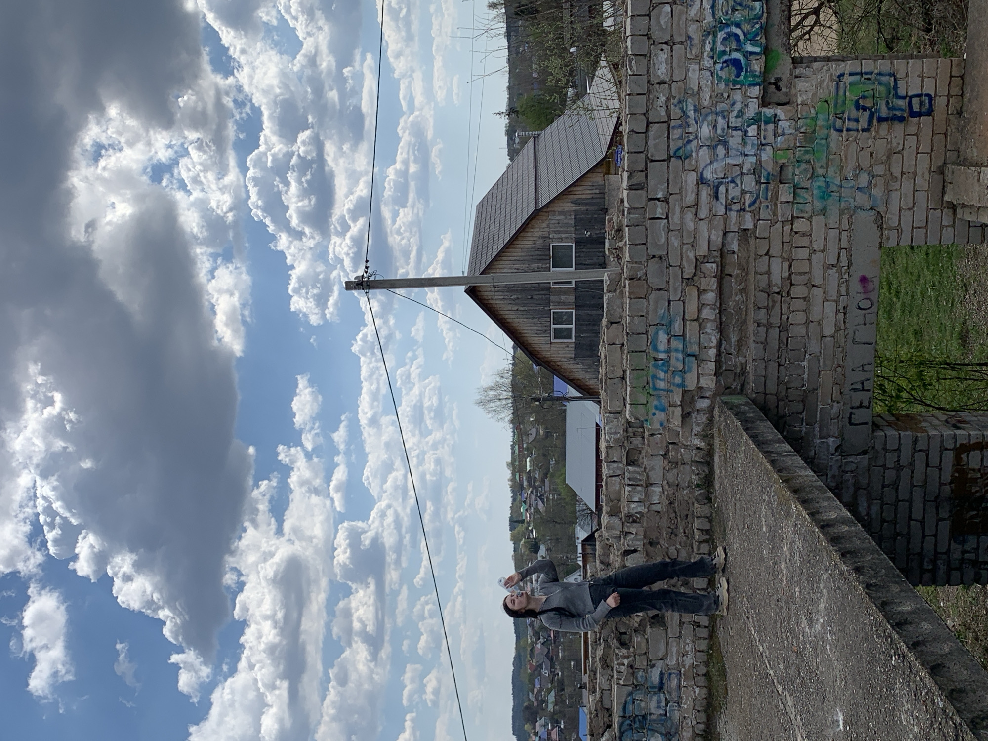 A full shot depicts a person standing on a concrete structure with a brick wall adjacent to it. The brick wall has graffiti on it. The person is dressed in dark pants and a gray top and appears to be drinking from a water bottle. A wooden house with a metal roof is visible in the background, along with an electrical pole and wires. Beyond the house, a cityscape extends into the distance under a sky with scattered clouds. The image location is Фоки, Russia.