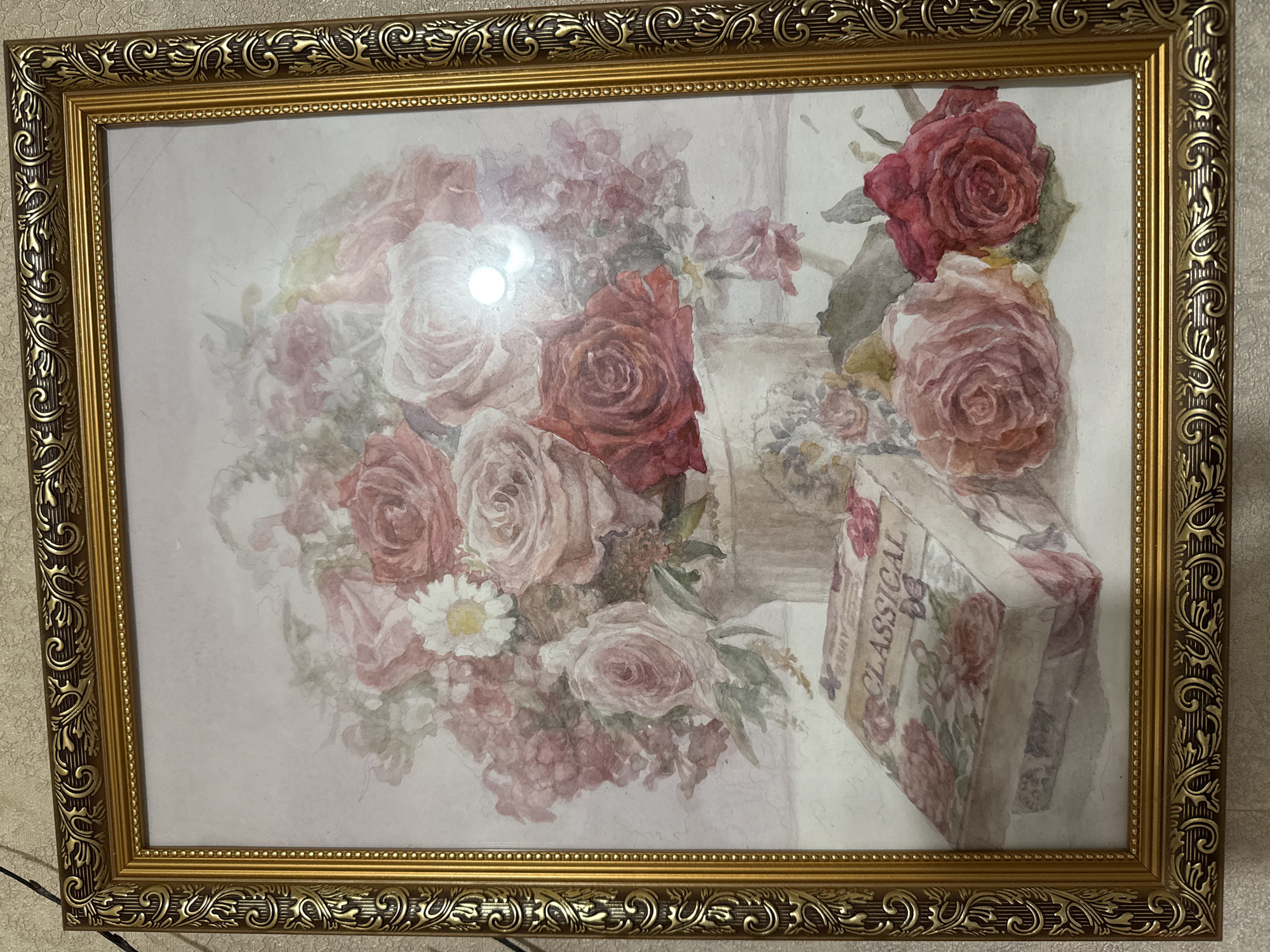 A framed floral still-life painting hangs on a light-colored textured wall. The frame is ornate gold. The painting depicts a bouquet of pink and red roses in a vase, with a small box labeled "CLASSICAL" sitting next to it. The light suggests an indoor setting. The location is not evident from the image.