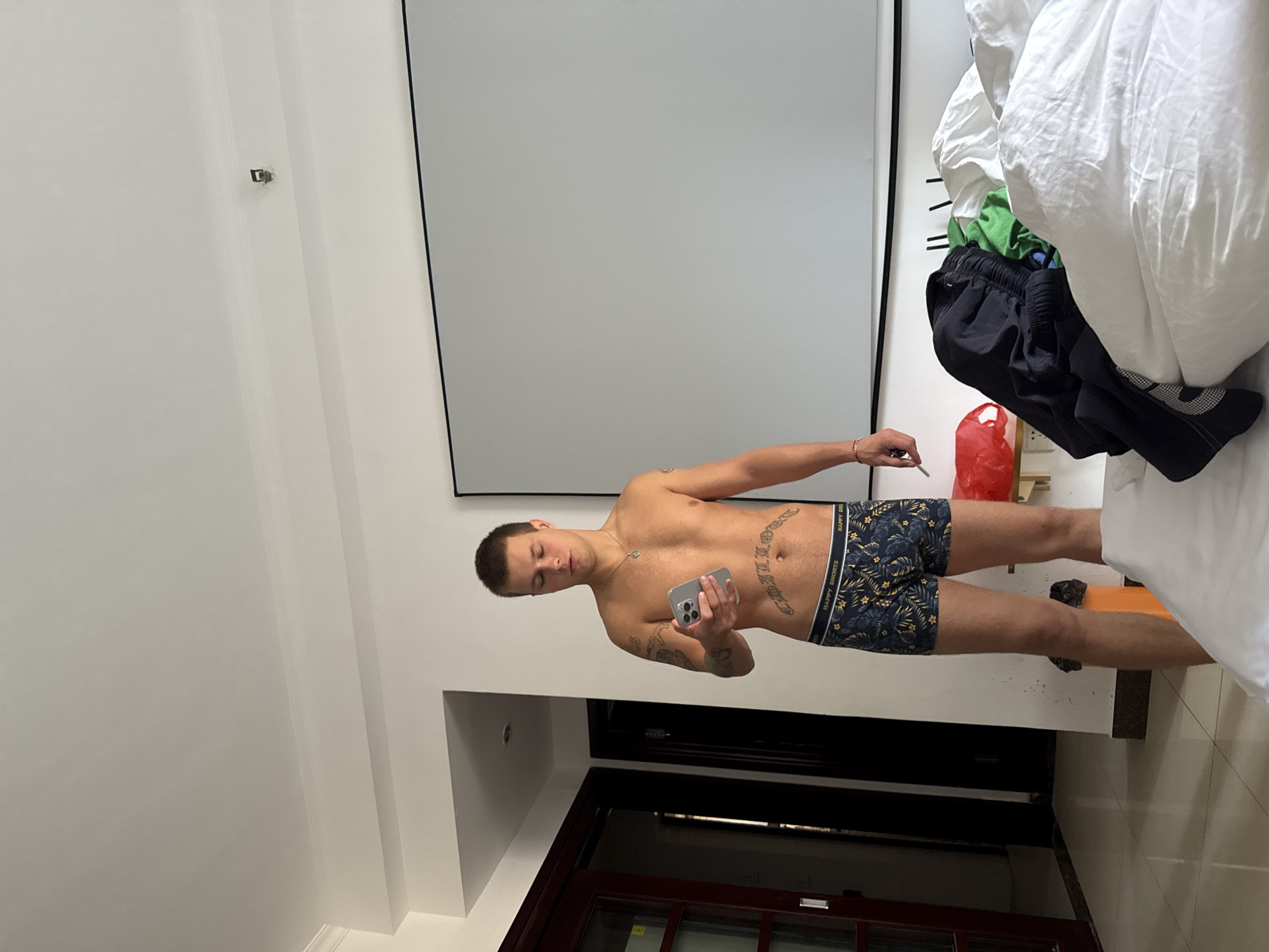 A shirtless, fair-skinned man with tattoos is taking a selfie in a room in Haizhu District, China. He is wearing patterned underwear and holding a smartphone. The room features white walls, a large mirror or screen, a bed with white linens, and a dark wood-framed door. Clothes and a red plastic bag are piled near the bed.