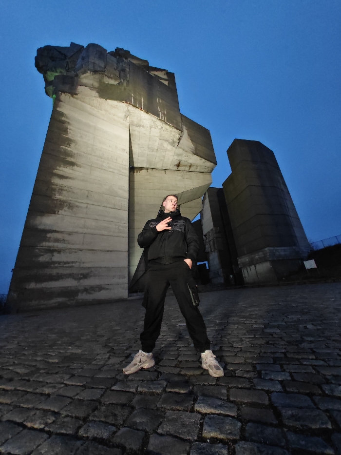 An adult male stands centrally in the foreground, facing slightly left of the camera against the backdrop of the Monument to 1300 Years of Bulgaria in Shumen, Bulgaria. The time of day appears to be twilight or early night, characterized by a deep blue, clear sky.
The subject is dressed in dark attire, consisting of a black padded jacket over a dark garment with a subtle pattern on the chest, and dark cargo-style pants. His footwear includes light-colored athletic shoes. The individual's left hand is raised to chest level, forming a gesture with the thumb and pinky finger extended, while his right hand rests inside his pants pocket. His gaze is directed upwards and slightly to his left.
The monumental concrete structure behind the individual exhibits a Brutalist architectural style, composed of large, angular blocks. Portions of the monument are illuminated by artificial lighting, displaying green and white hues on the concrete surfaces. Wear and discoloration are visible on the concrete. To the far right, a more columnar section of the monument stands against the sky. The ground beneath the subject is paved with dark, irregularly shaped cobblestones. The overall scene is lit by ambient twilight and artificial monument lighting.