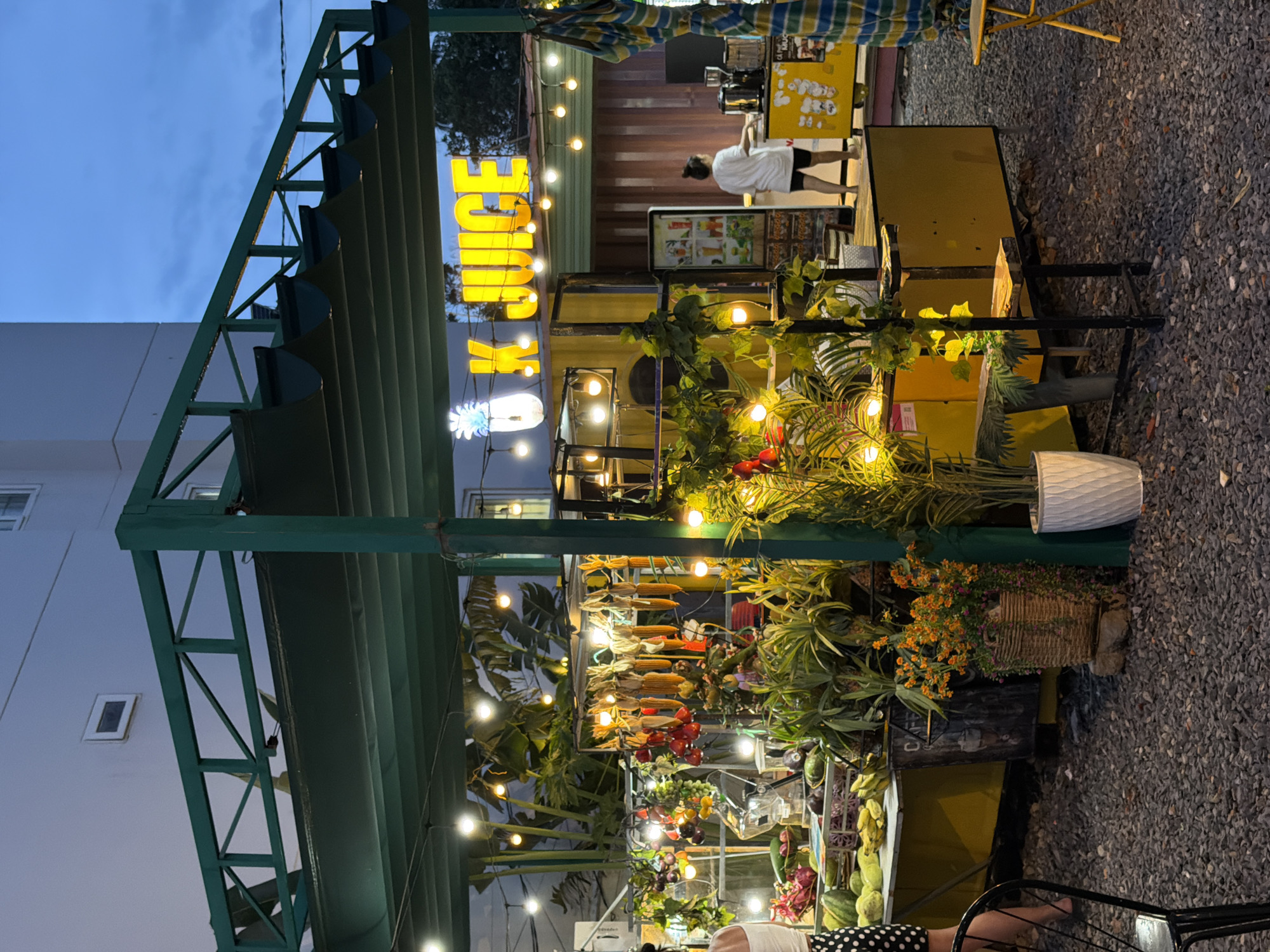 This image captures an outdoor juice stand in Nha Trang, Vietnam. The stand features a bright yellow counter adorned with string lights and a prominent illuminated sign that reads "K. JUICE" with a pineapple graphic. Various fresh fruits and vegetables are displayed on the counter, including corn, bananas, and leafy greens. A woman in a white shirt and black shorts is seen behind the counter, appearing to operate the juice stand. The ground is covered in gravel, and potted plants add a natural element to the scene. In the background, a building suggests an urban setting, while the overall ambiance is that of a casual, evening marketplace.