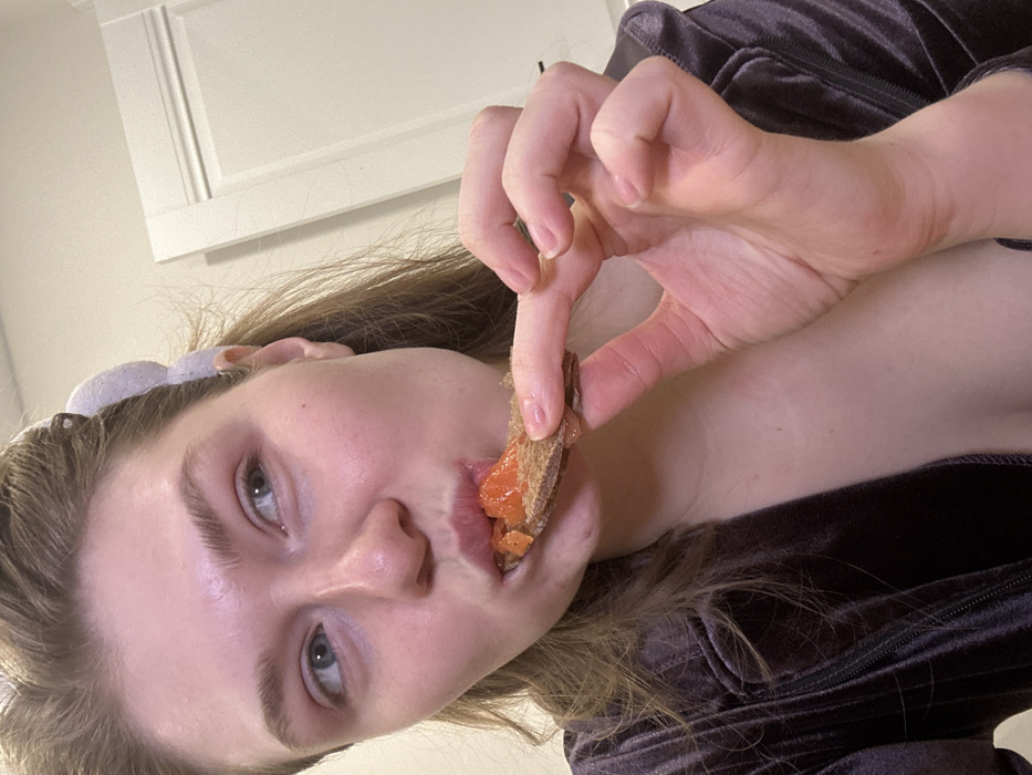 A woman in a grey velvet top holds a small piece of bread topped with an orange substance to her lips. Her left hand forms a loose "OK" gesture around the snack. The woman's pale skin is contrasted by her light brown hair and grey eyes. A white headband sits atop her head. The background features a white wall with a framed, paneled section. The photo is taken from a slightly upward angle.