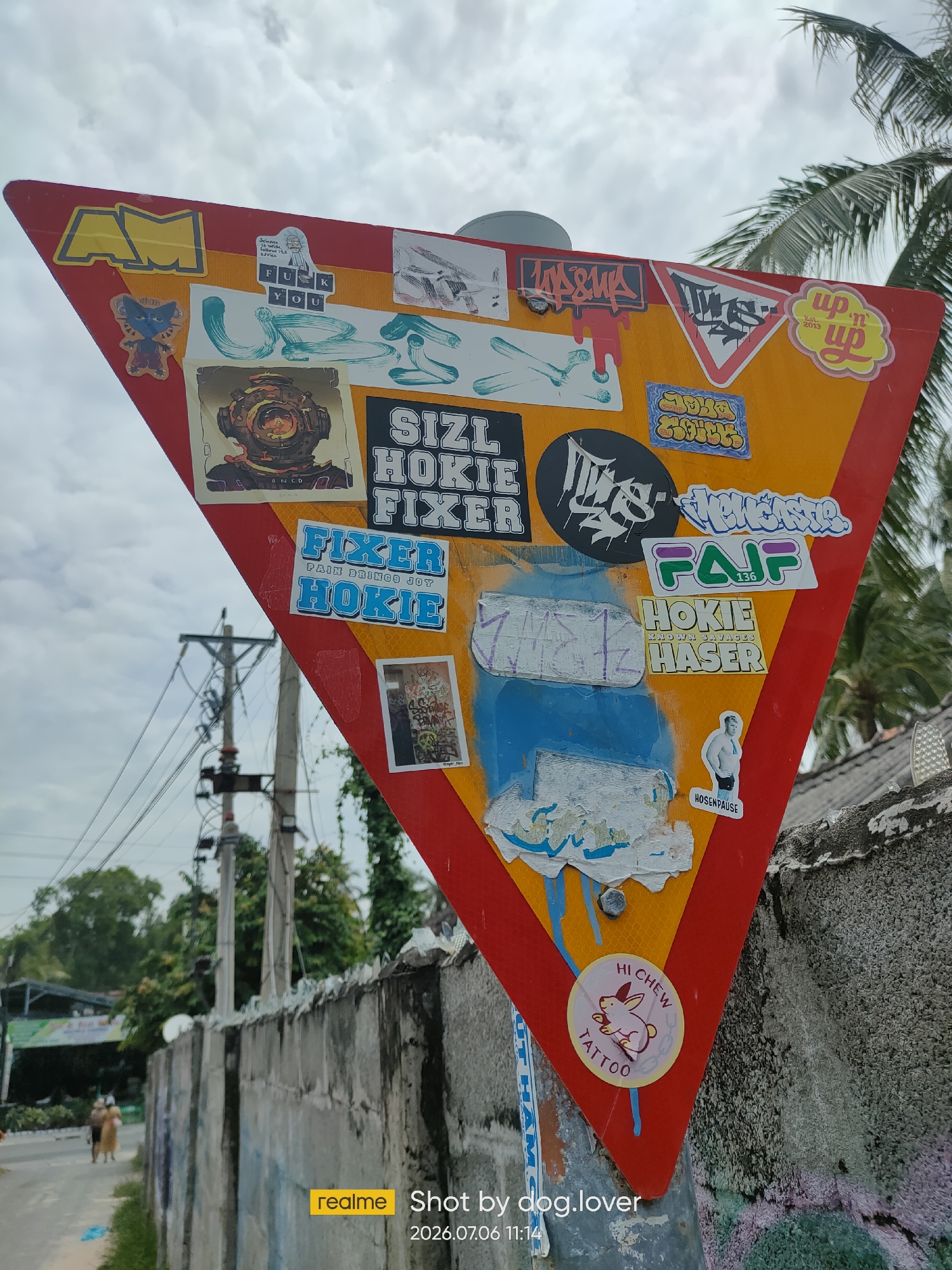 A triangular yield sign, predominantly yellow with a red border, is heavily adorned with various stickers. The sign is located on a pole next to a concrete wall, with a palm tree visible in the upper right corner. In the background, a street runs alongside the wall, with two individuals walking away from the camera. A utility pole with numerous wires is situated behind the yield sign. The sky is overcast with clouds. The bottom right of the image shows a watermark indicating "realme Shot by dog.lover 2026.07.06 11:14".
