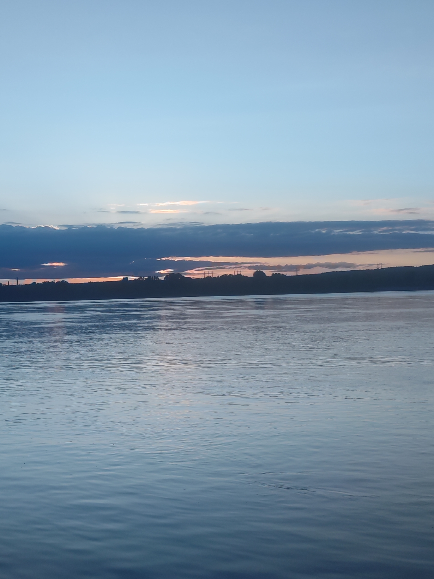 The image presents a serene twilight landscape over a wide body of water, likely a large river or lake, set against the backdrop of a transitioning sky. The time of day appears to be either sunrise or sunset, indicated by the soft orange, pink, and yellow light breaking through gaps in a prominent band of dark, horizontal clouds positioned just above the distant horizon. The upper sky is a clear, light blue, gradually darkening towards the cloud layer.
The lower half of the image is dominated by the calm water, displaying gentle ripples and reflecting the muted blue and grey tones of the sky. Its surface is undisturbed, contributing to a sense of tranquility. A dark, indistinct silhouette of land stretches across the middle distance, separating the water from the sky. This shoreline appears densely vegetated, possibly tree-lined, and in places, faint outlines of human-made structures, such as power line towers and a distant tall mast or smokestack, are discernible within the silhouette.
The scene is devoid of people or explicit activities, focusing entirely on the natural beauty of the landscape at dusk or dawn. The weather appears calm and clear, with the clouds adding a dramatic element to the sky. This tranquil vista is situated in the context of Noviy, Russia, offering a peaceful view of the region's expansive waters. No text is visible in the media file.