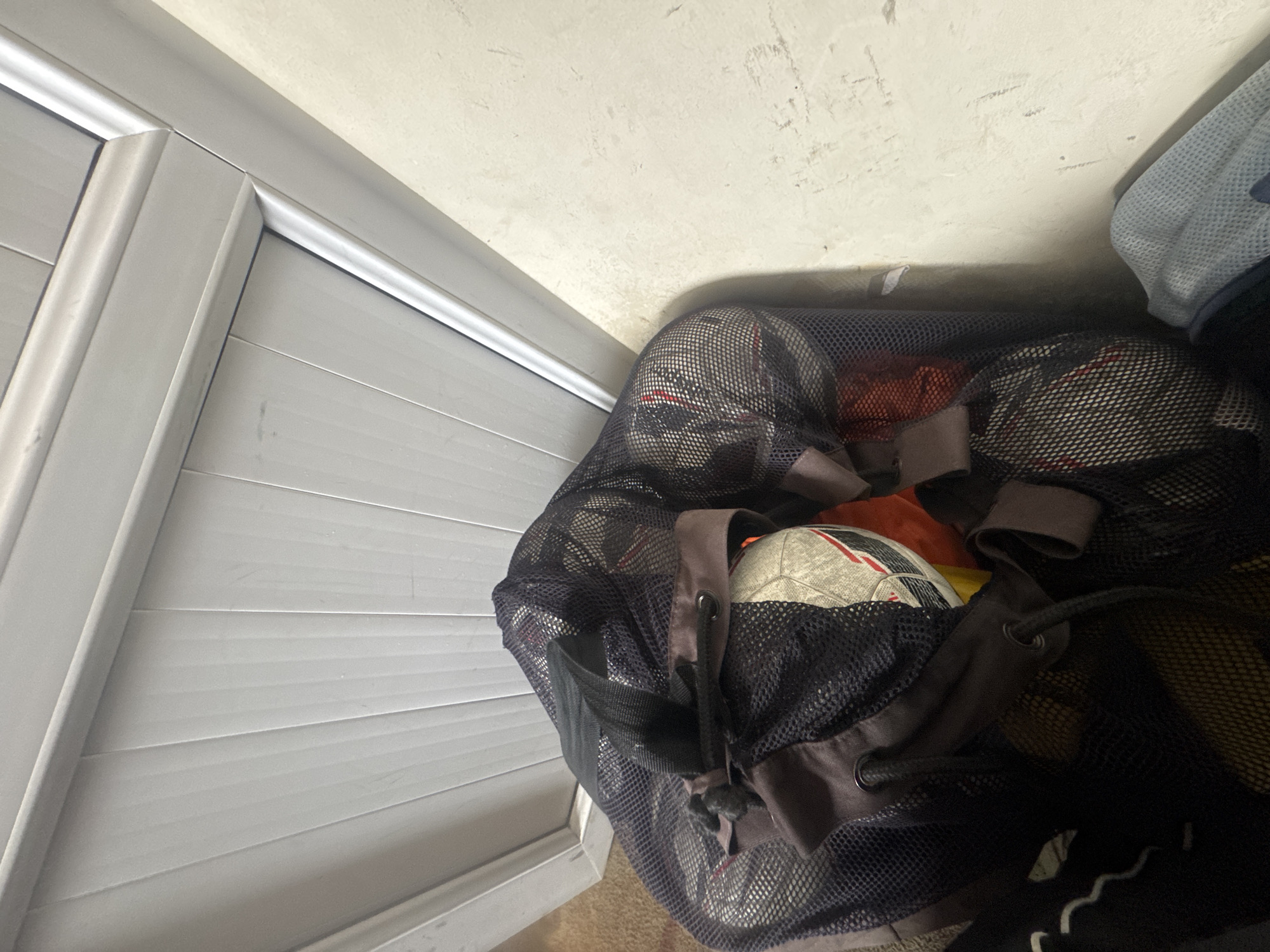 The image depicts a mesh bag filled with soccer balls and other items, positioned against a white wall and a white louvered door. The bag is dark and the contents are partially visible, including the patterned surfaces of the soccer balls. The environment appears to be indoors. The location of Charentsavan, Armenia is not verifiable based on visual information within the image.