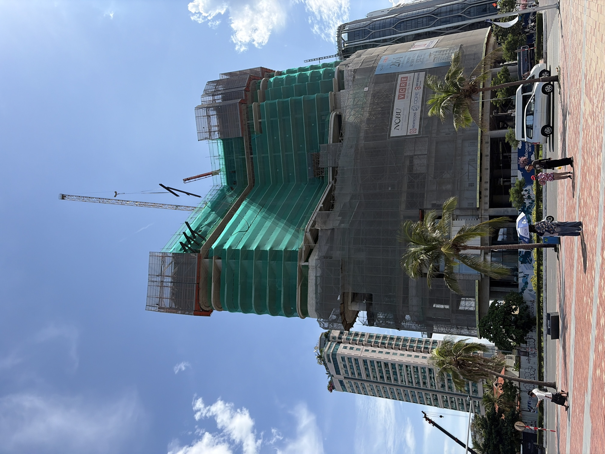 The image depicts a construction site in Đà Nẵng, Vietnam, featuring a partially built skyscraper covered in green netting. In the foreground, a sidewalk paved with red and beige bricks leads to a street. Three people are visible on the sidewalk: one person walking with a bag, and two people standing near each other. One of the standing individuals holds a blue umbrella. Across the street, several vehicles are present, including a white van, a bus, and what appears to be a white car. Palm trees are scattered throughout the scene, adding to the tropical atmosphere. To the left of the main construction site, another taller building stands, and a construction crane is visible in the background. The sky is blue with scattered white clouds, suggesting a clear day.