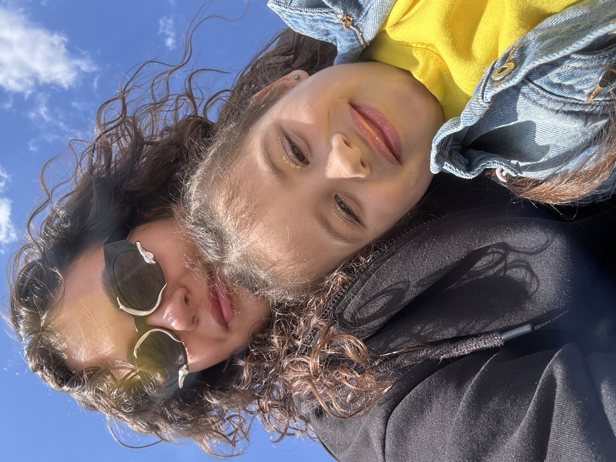 Two individuals are visible in this rotated selfie taken outdoors under a bright blue sky with sparse white clouds. The background indicates daylight conditions.
The individual at the bottom wears sunglasses with a distinctive silver embellishment and a black hooded sweatshirt with visible zipper details. Curly brown hair surrounds their face.
The individual at the top wears a yellow top under a denim jacket. They have brown hair and visible makeup, including glitter eyeshadow and glossy lips. They are smiling slightly.
The close proximity of the individuals suggests they are posing together. There are no indicators of Prijevor, Montenegro.