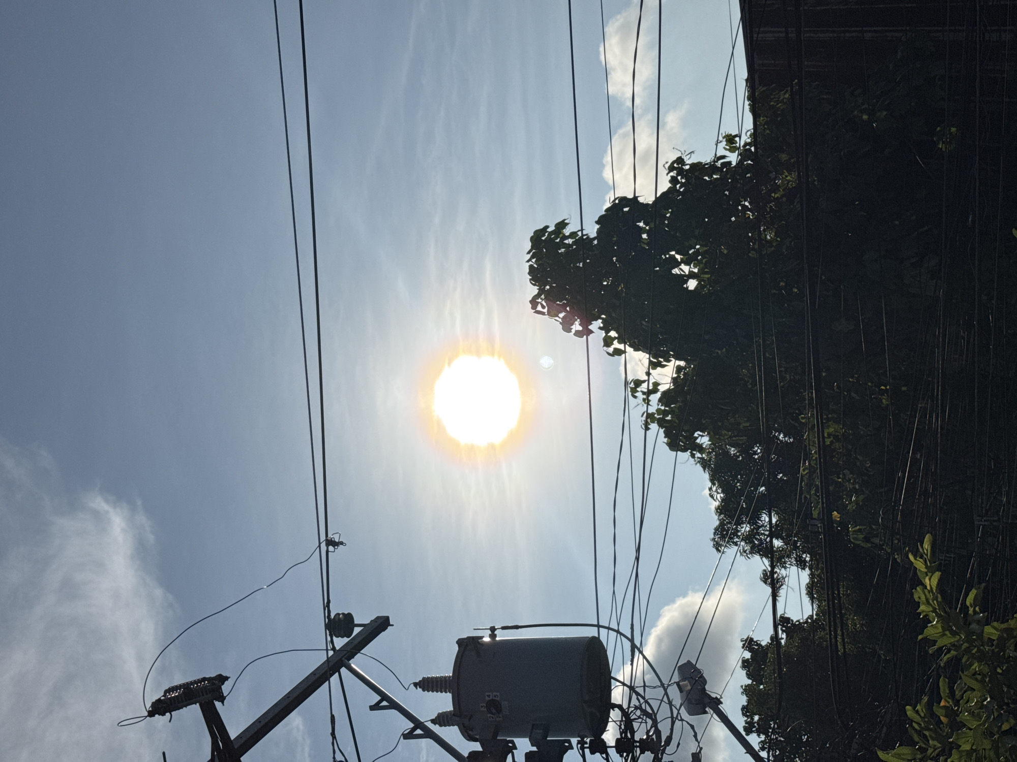 The image depicts a view looking upwards in an urban environment, likely in Davao City, Philippines. The sky is light blue with scattered clouds and the sun is visible, brightly shining through the haze. Overhead power lines crisscross the frame, converging toward a utility pole and transformer assembly. Trees with dense green foliage occupy the right side of the image, obscuring parts of the sky. The building can be seen at the upper right corner of the image.