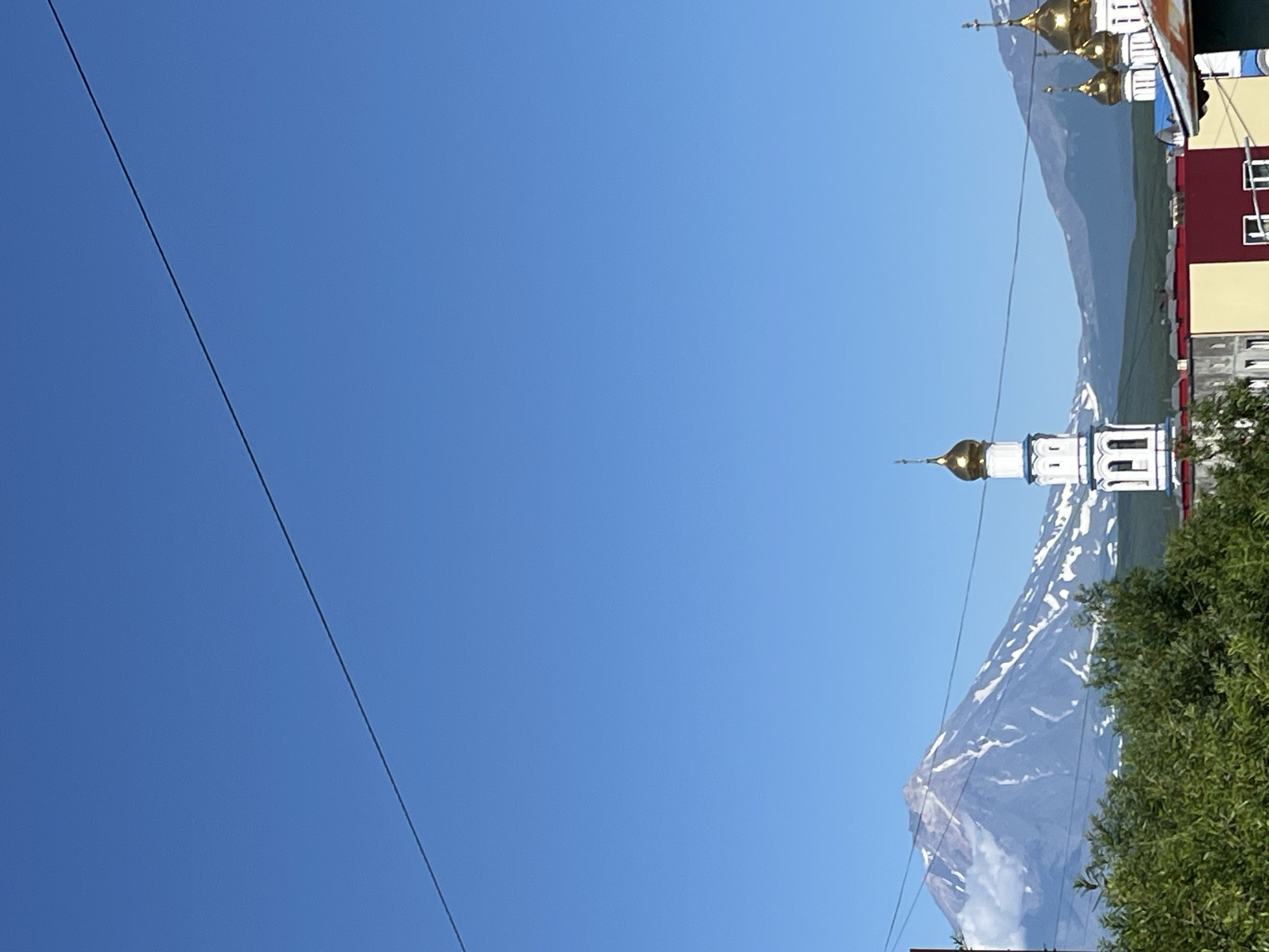 The image captures a view of Petropavlovsk-Kamchatsky, Russia, featuring a prominent snow-capped volcano in the background under a clear blue sky. In the foreground, a white and blue bell tower with a golden dome rises, characteristic of Orthodox church architecture, alongside other buildings with red and yellow facades. Green trees partially obscure the lower portion of the scene. A thin wire stretches across the sky, a common element in urban landscapes. The overall impression is one of a city nestled within a dramatic natural landscape.