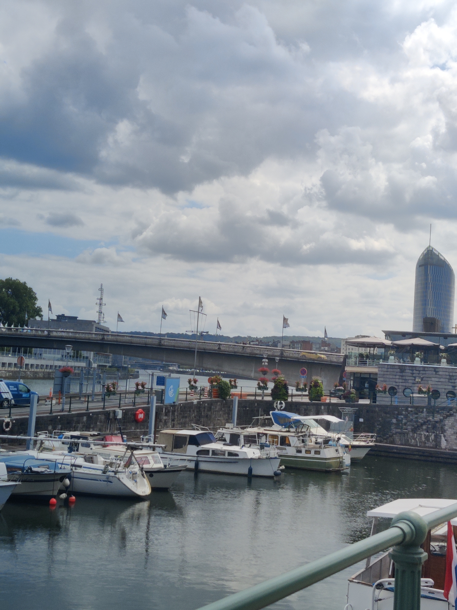 The image shows a waterfront scene in the city of Liège, Belgium, likely near the Quai de la Batte. The sky is overcast with dramatic clouds. In the foreground, several boats are docked in a marina. These boats are varied in size and style, including sailboats and motorboats, some covered with tarpaulins. The water in the marina reflects the boats and the sky.
In the mid-ground, a wide bridge spans the water, with a road carrying traffic. Beyond the bridge, buildings line the waterfront, including what appears to be a modern, tall glass building with a distinctive curved shape on the right side of the frame. This building is likely the assumed location of the "Louveciennes" building or another prominent skyscraper along the Meuse River in Liège. Flags are visible on poles along the bridge and waterfront.
The overall atmosphere is one of a typical day in a European city's harbor, with commercial activity implied by the presence of boats and infrastructure. There are no visible people in the image. The scene is tranquil, with the primary focus on the boats, the bridge, and the urban skyline.