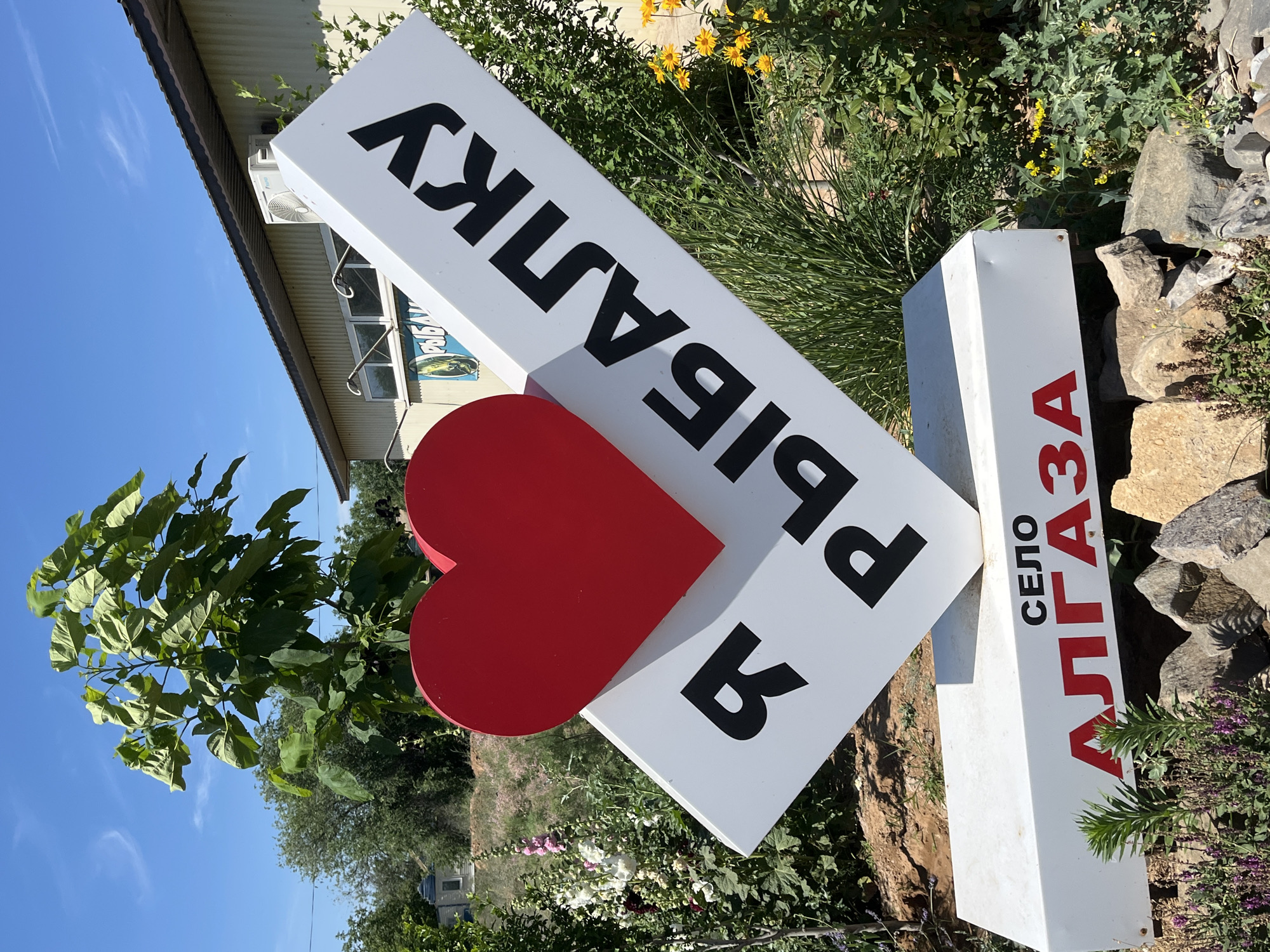 A vertical outdoor photograph shows a white sign with black Cyrillic lettering and a red heart. The sign is tilted, and its text reads "Я ЛЮБЛЮ РЫБАЛКУ" which translates to "I love fishing." A second, smaller white sign is to the right of the first, with red lettering that says "СЕЛО АЛГАЗА," identifying the location as the village of Algaza. The setting appears to be a rural area in Algaza, Russia, with lush green foliage, flowering plants, and rough stones visible in the background. The sky is a clear, bright blue, suggesting a sunny day. The composition, with the prominently displayed "I love fishing" sign and the village name, implies this is a local landmark or attraction for visitors and residents. There are no people or specific activities depicted; the focus is solely on the signs and their surroundings.