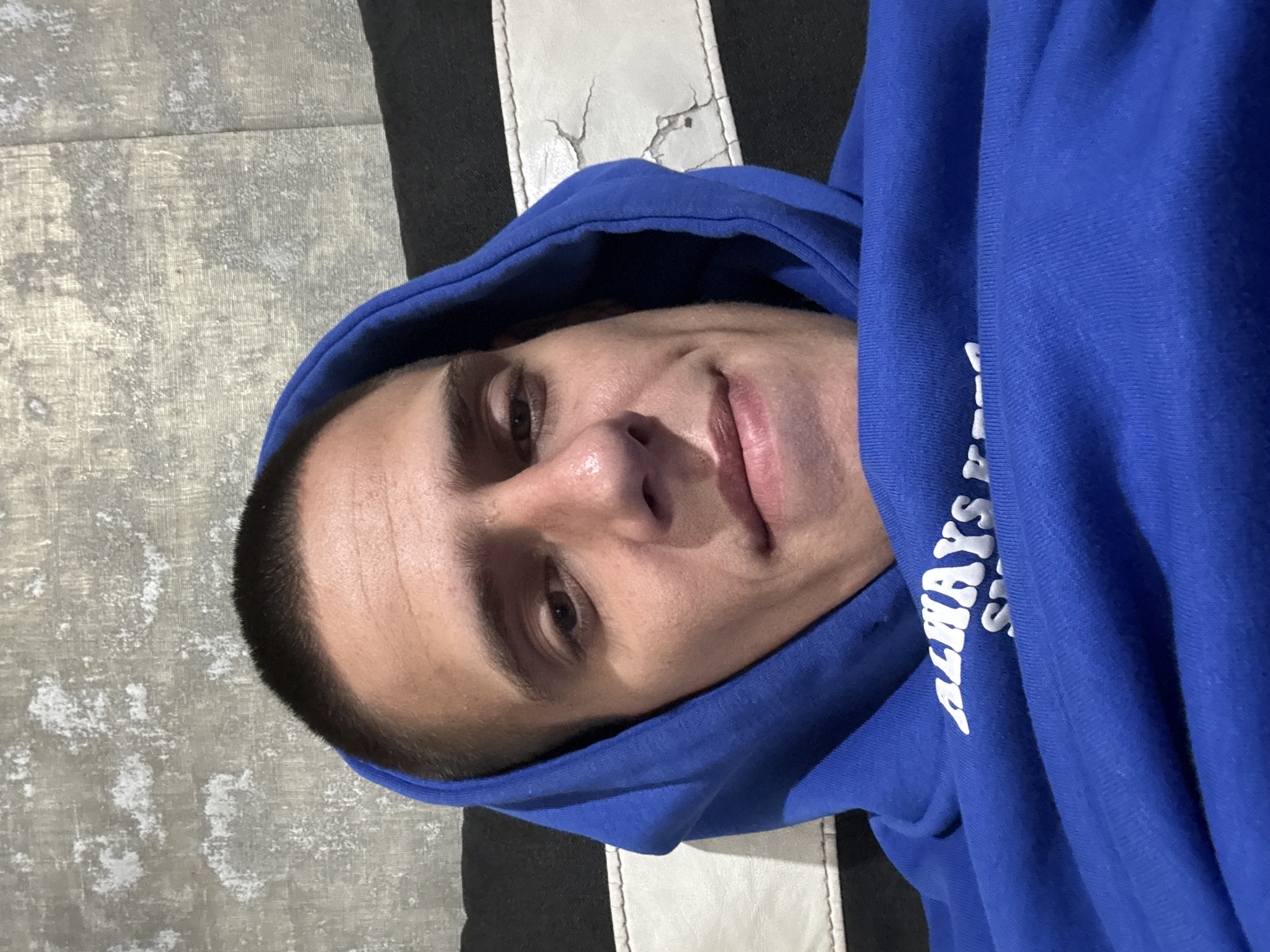 A close-up, low-angle selfie shows a young Caucasian male with short dark hair, wearing a bright blue hoodie. He is lying down, possibly on a bed or sofa, with a textured grey wall and a striped black and white surface visible in the background. The hoodie has white text printed on it that is partially visible, reading "ALWAYS". The man's gaze is directed towards the camera, and he has a slight smile. The lighting appears to be artificial, coming from above and slightly to the front. The image was taken indoors, in a residential setting. There are no specific visual cues to indicate the location in Sofia, Bulgaria.